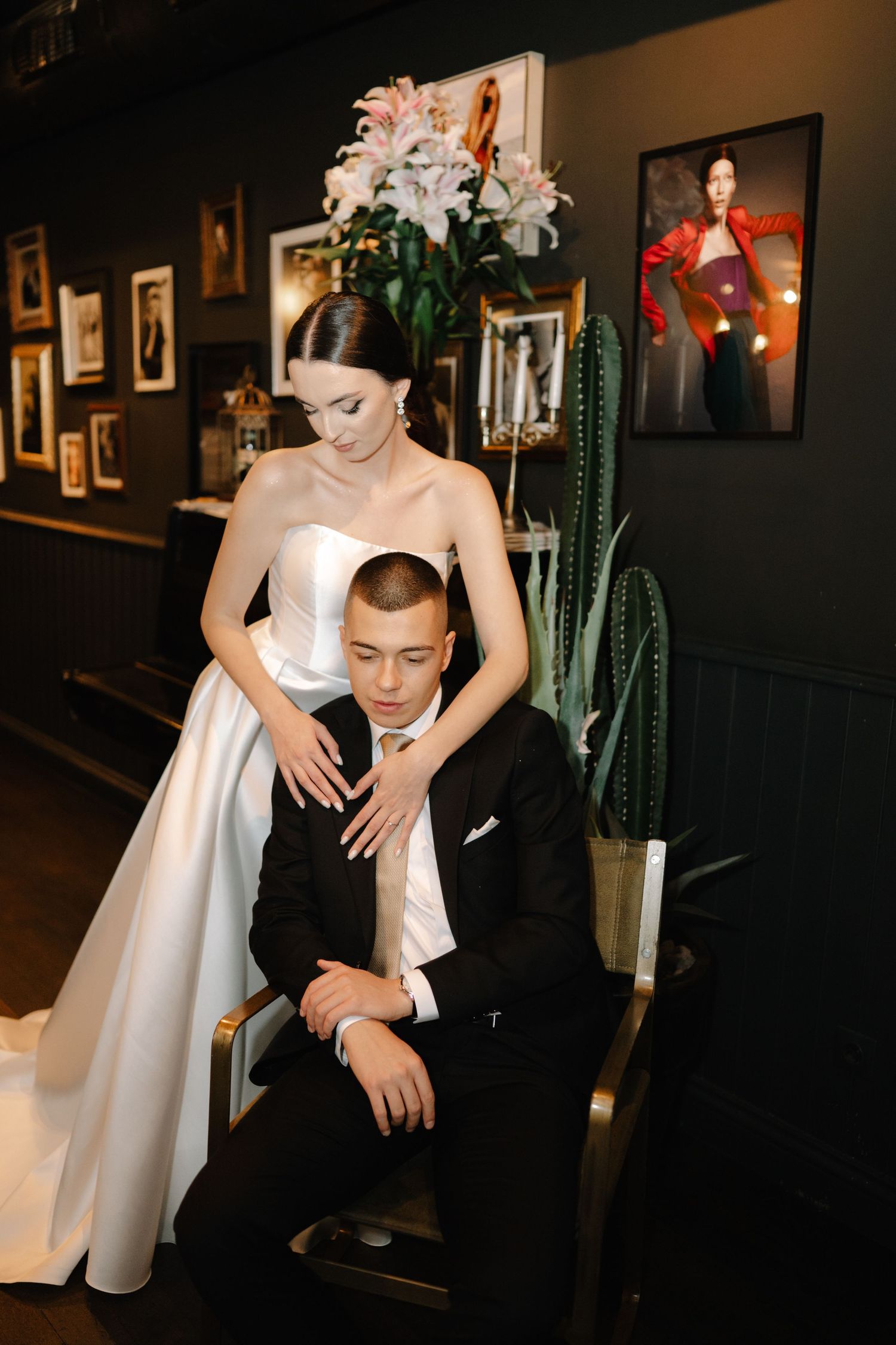 Elegant couple in wedding attire pose in a dark room with a cactus plant and framed artwork on the wall.