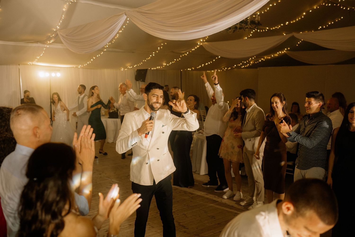 Groom in white jacket dancing energetically with guests at elegant wedding reception under fairy lights.