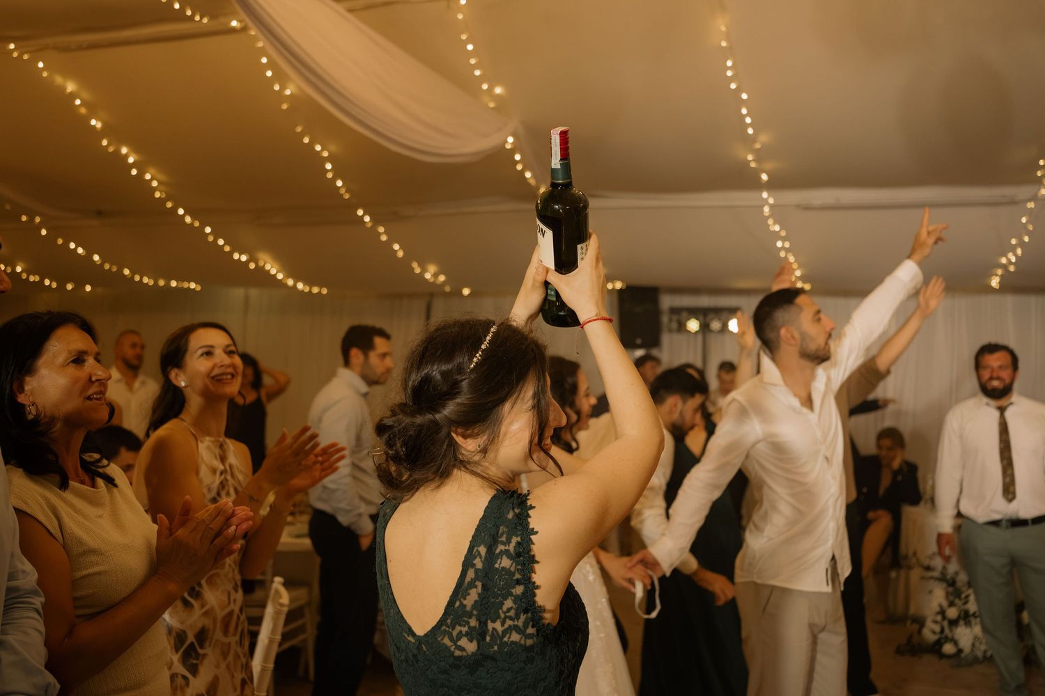 Guests celebrate at a wedding reception, dancing and raising a wine bottle under fairy lights in a decorated venue.