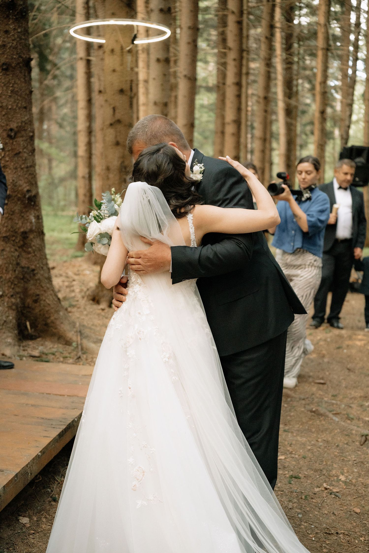 Bride and groom share an emotional embrace during outdoor forest wedding ceremony, bride holding white floral bouquet.