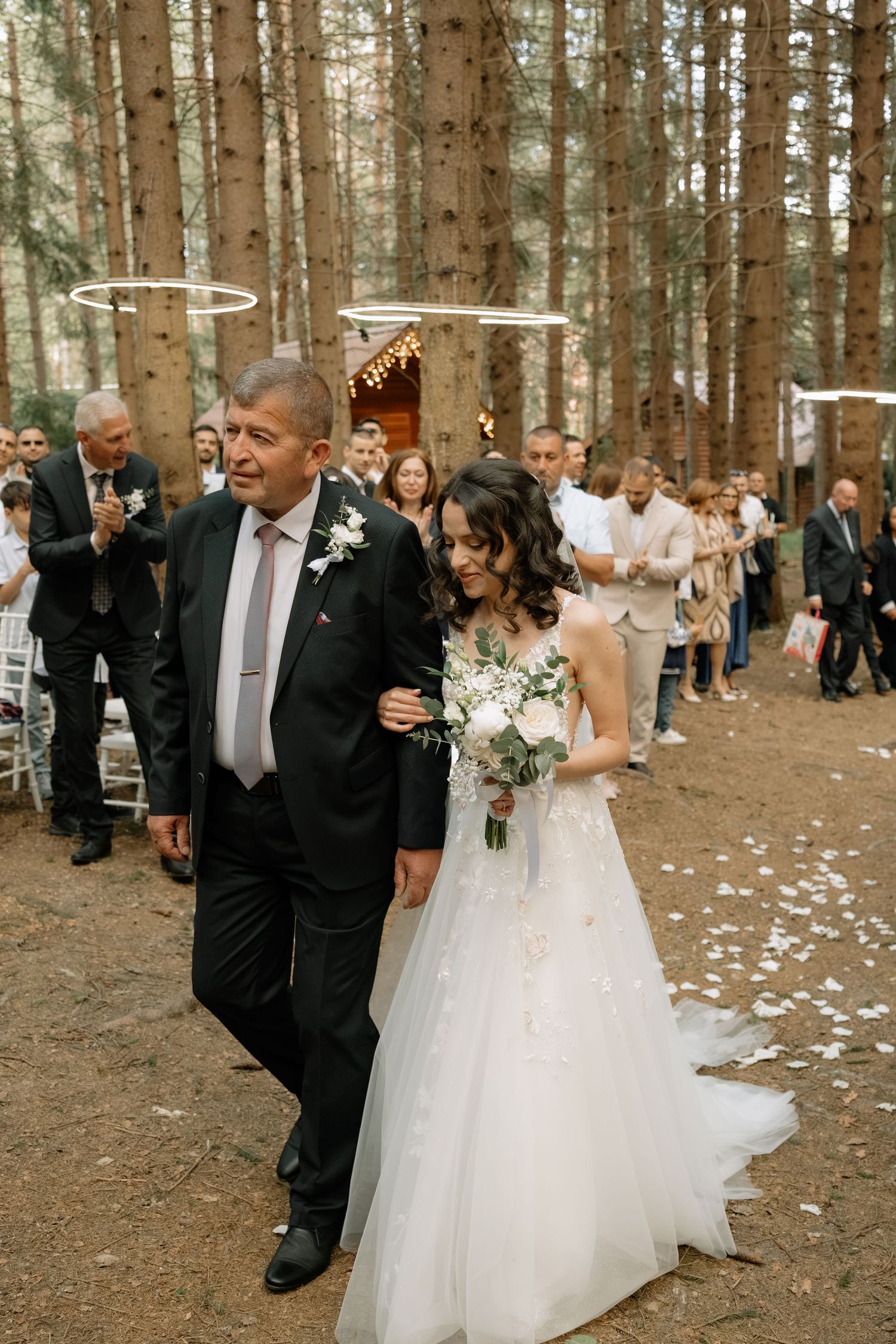 Bride in white gown walks with older man through forest wedding aisle, guests applauding, floral arch and pine trees backdrop.
