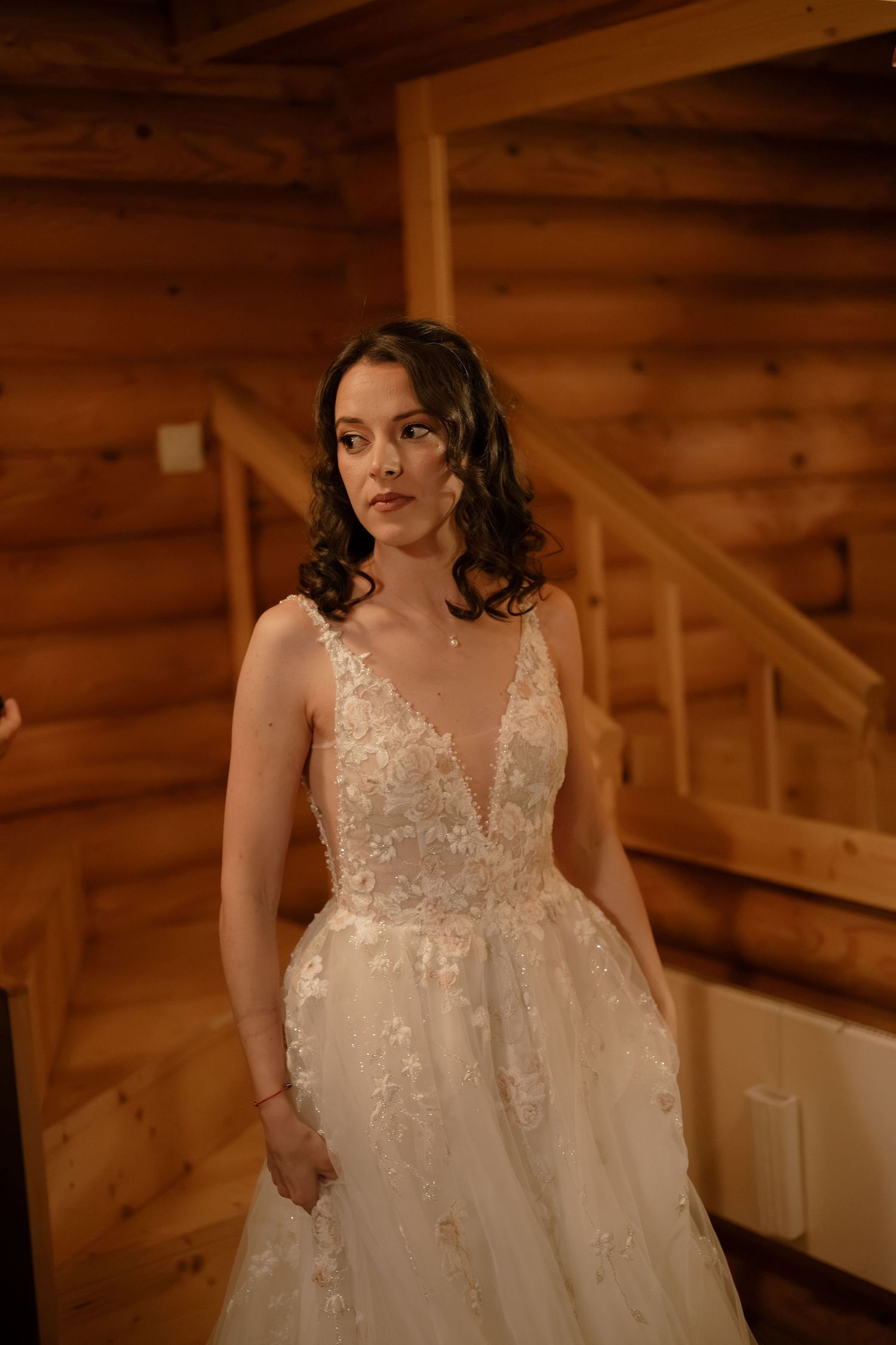 Bride in lace V-neck wedding gown poses in rustic log cabin with wooden staircase backdrop.