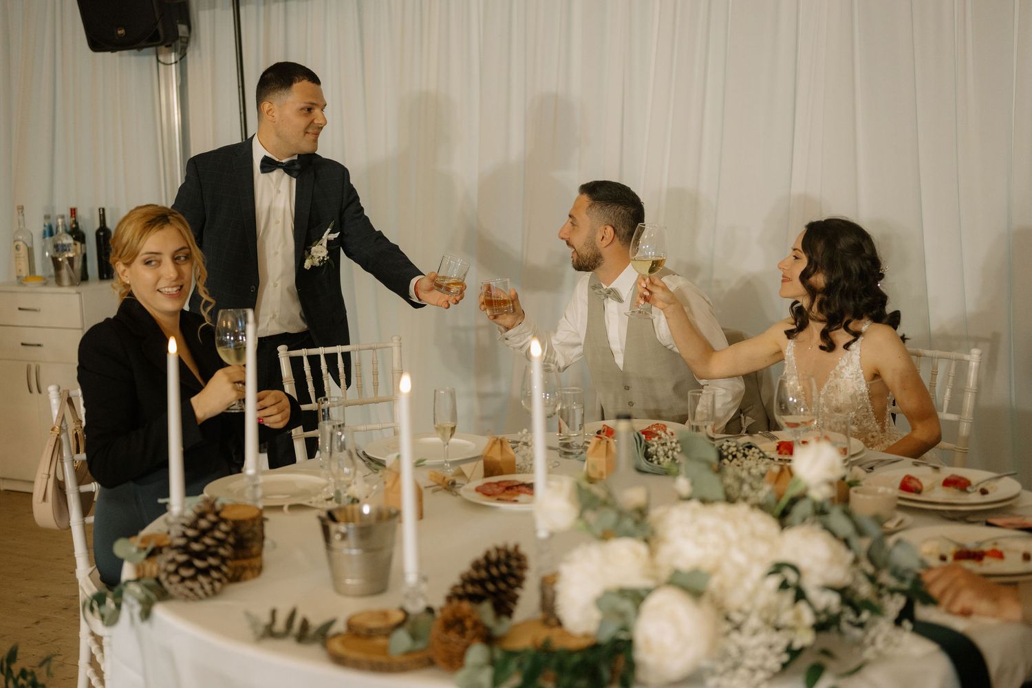 Wedding guests celebrating at elegant reception table with candles, pine cones, and white floral arrangements.