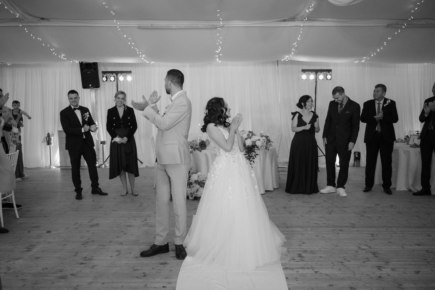Black and white photo of a bride and groom sharing their first dance surrounded by guests at an elegant wedding reception.