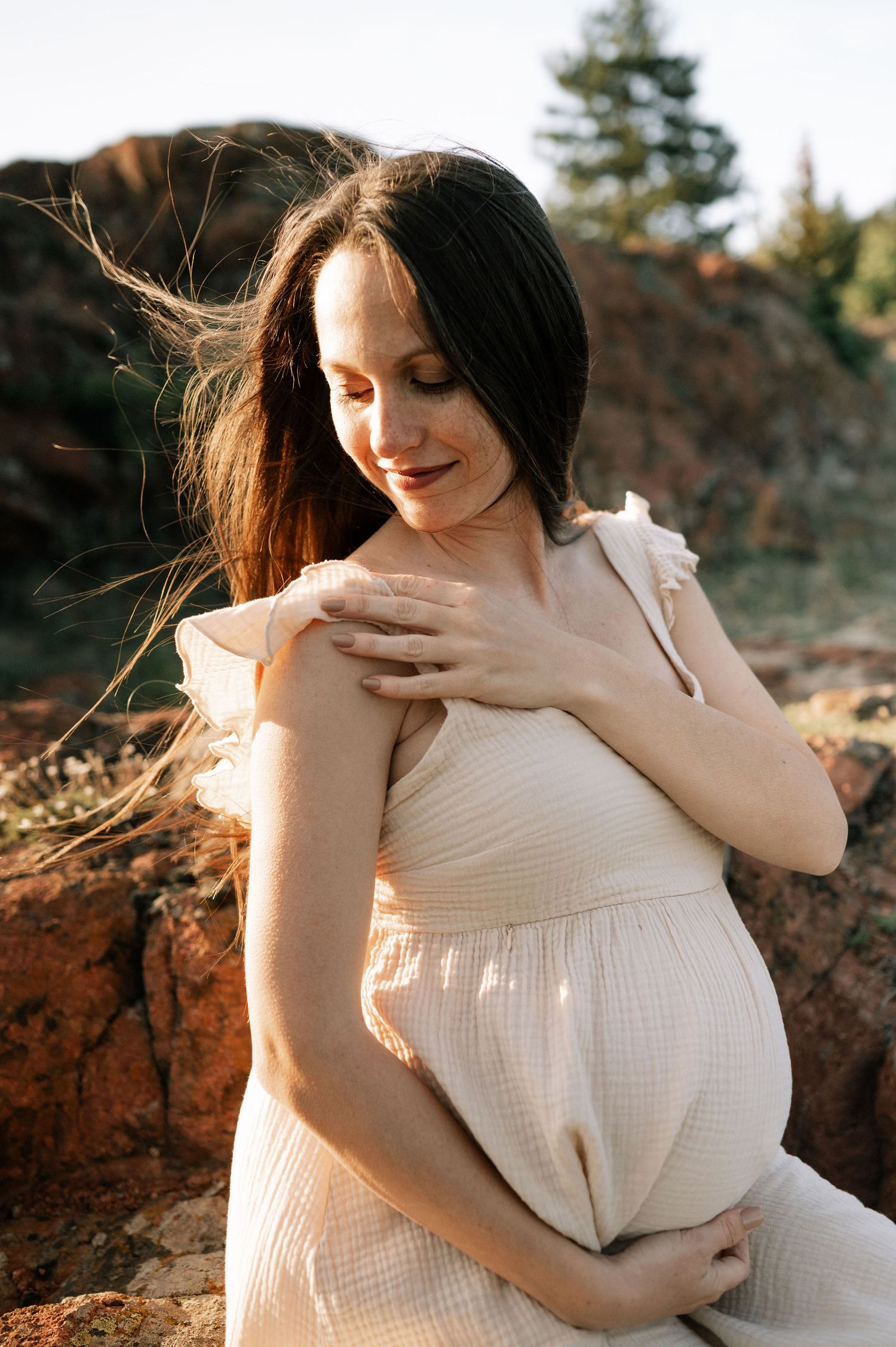 Pregnant woman in white dress touches her shoulder while posing against natural red rock backdrop at sunset.