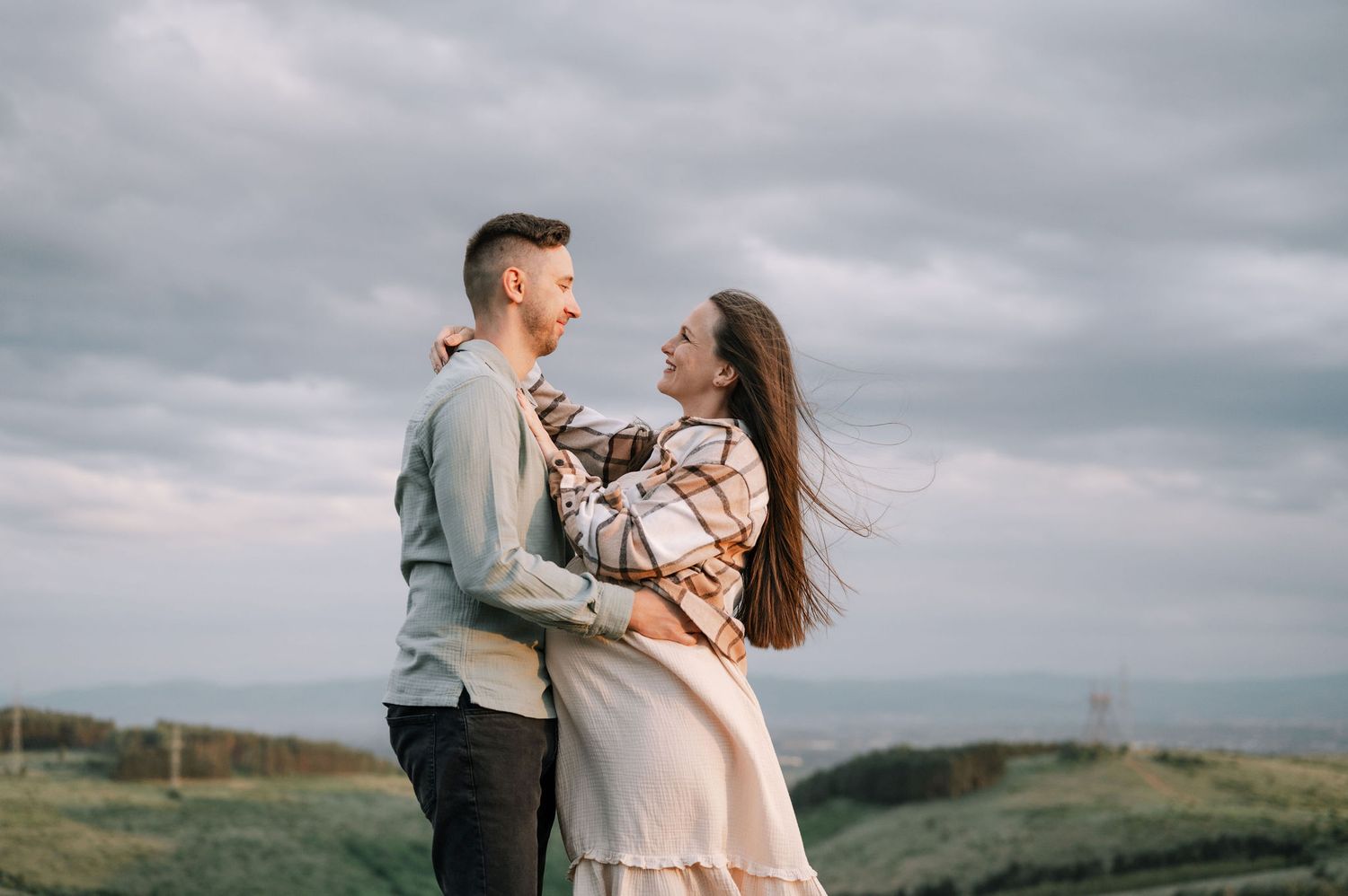 A couple embraces on a scenic hilltop overlooking a moody landscape during a cloudy afternoon.