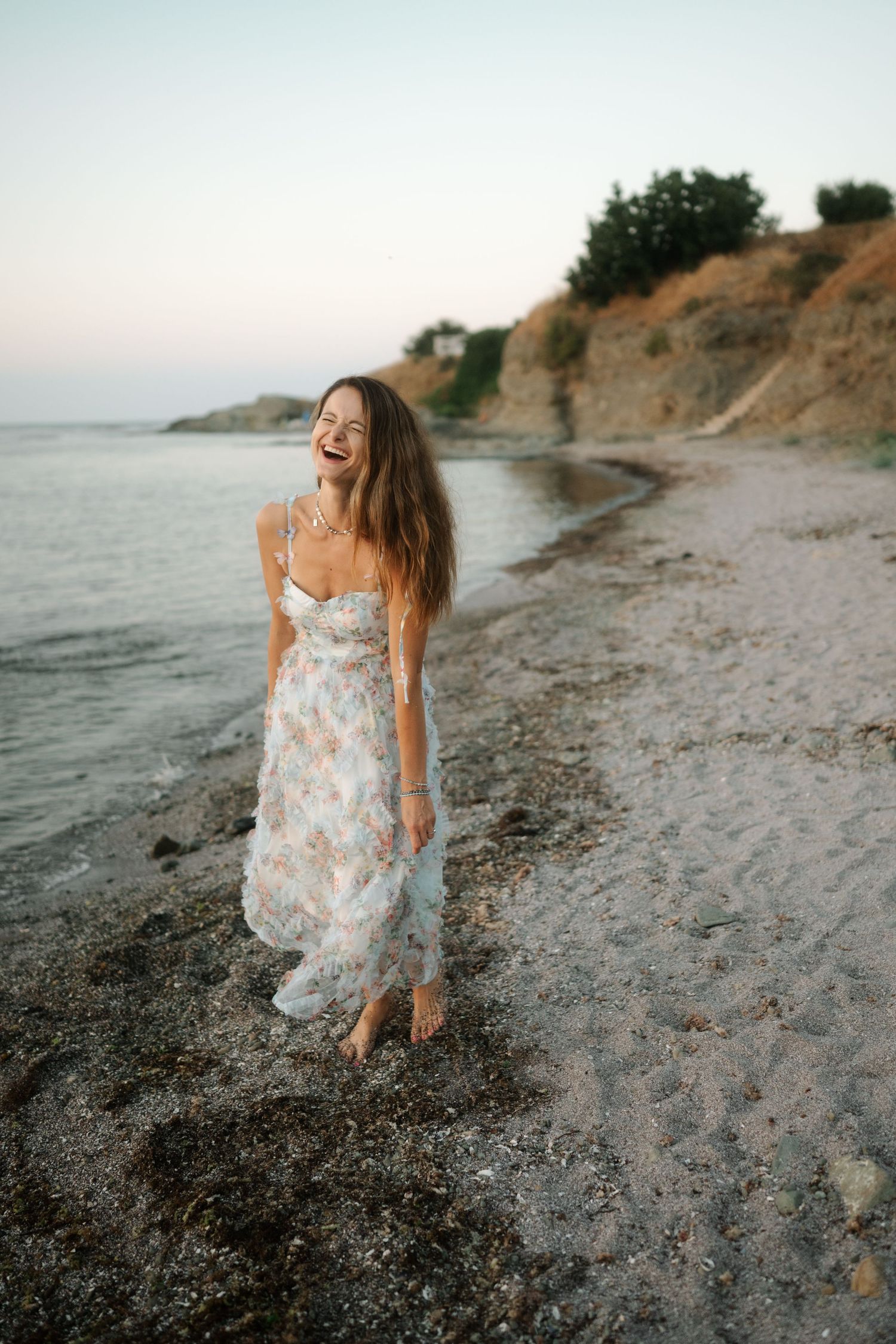A person in a floral dress runs joyfully along a rocky beach at sunset with cliffs in the background.