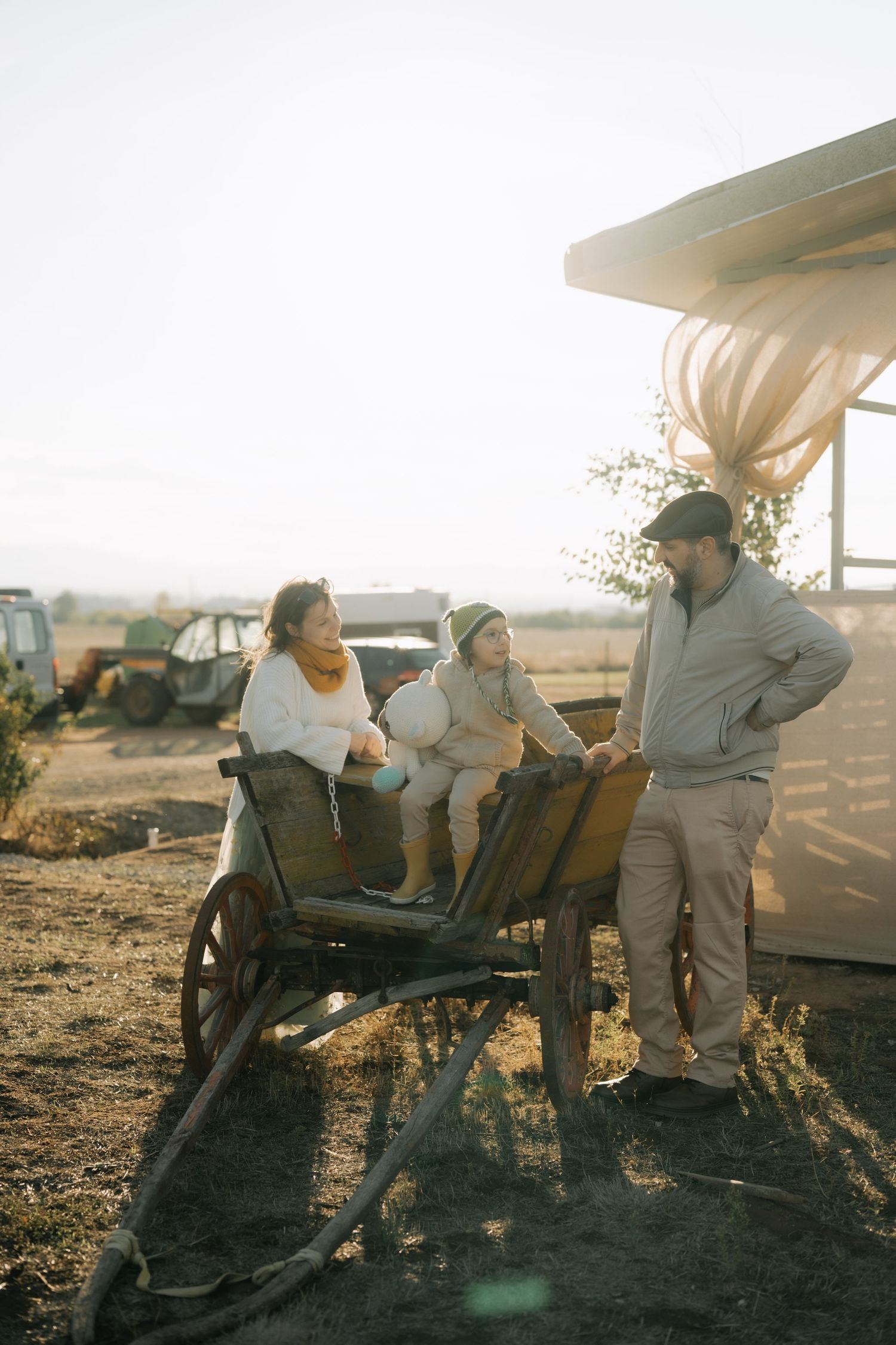 People gathered around an old wooden cart on a farm at sunset with equipment and buildings in the background.