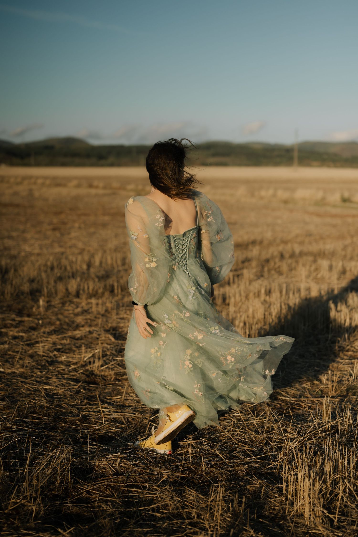 A figure in a flowing mint green dress twirls through a golden wheat field at sunset, creating a dreamy pastoral scene.