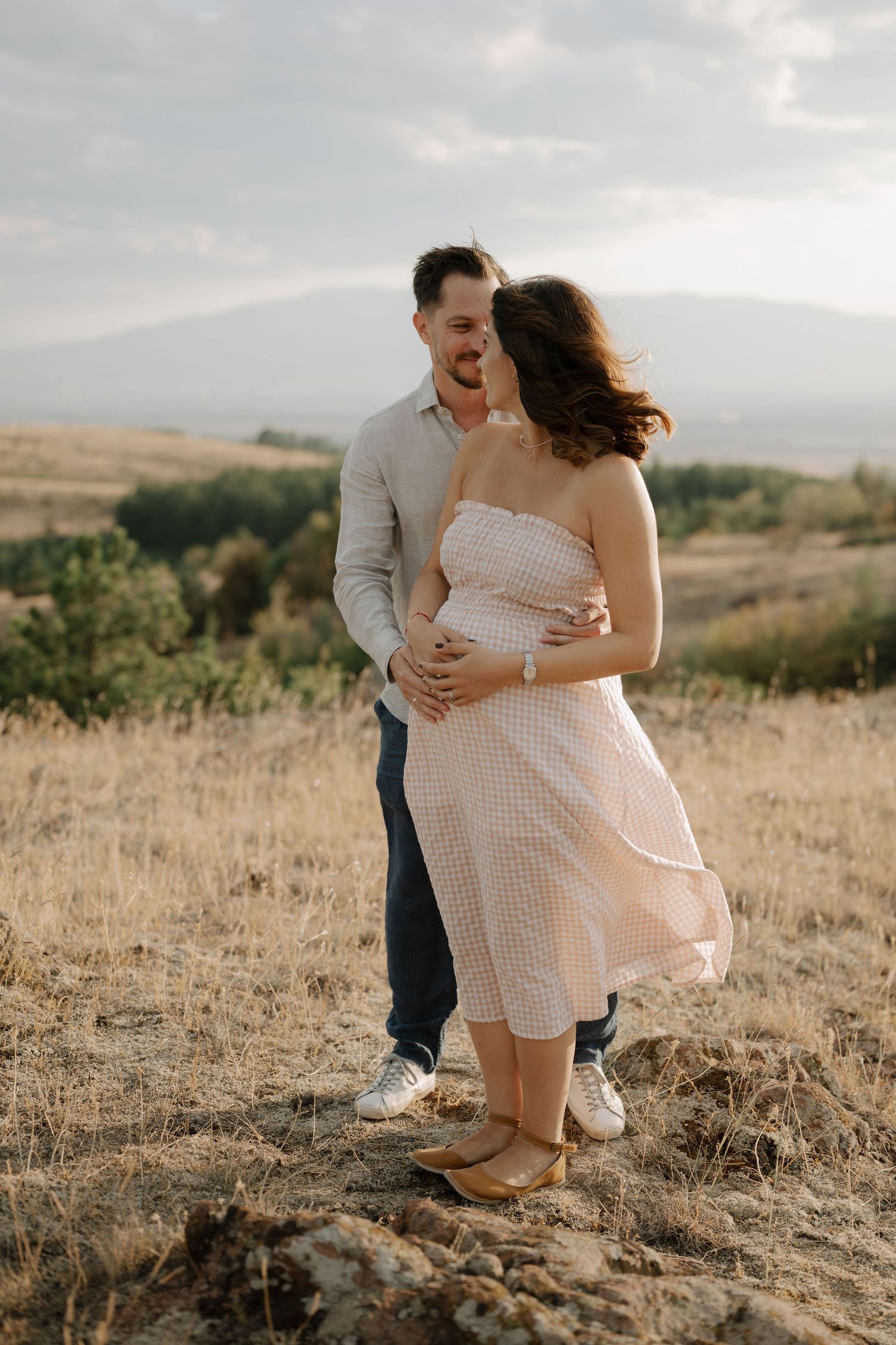 Romantic couple embraces in a sun-drenched field with rolling hills in the background during golden hour.