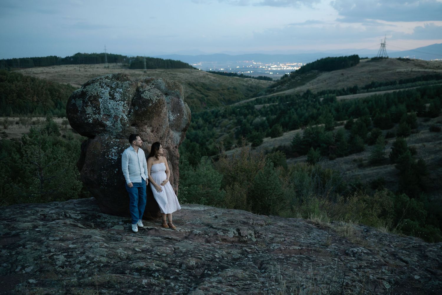 Two people stand on rocky terrain overlooking scenic mountain landscapes at sunset.
