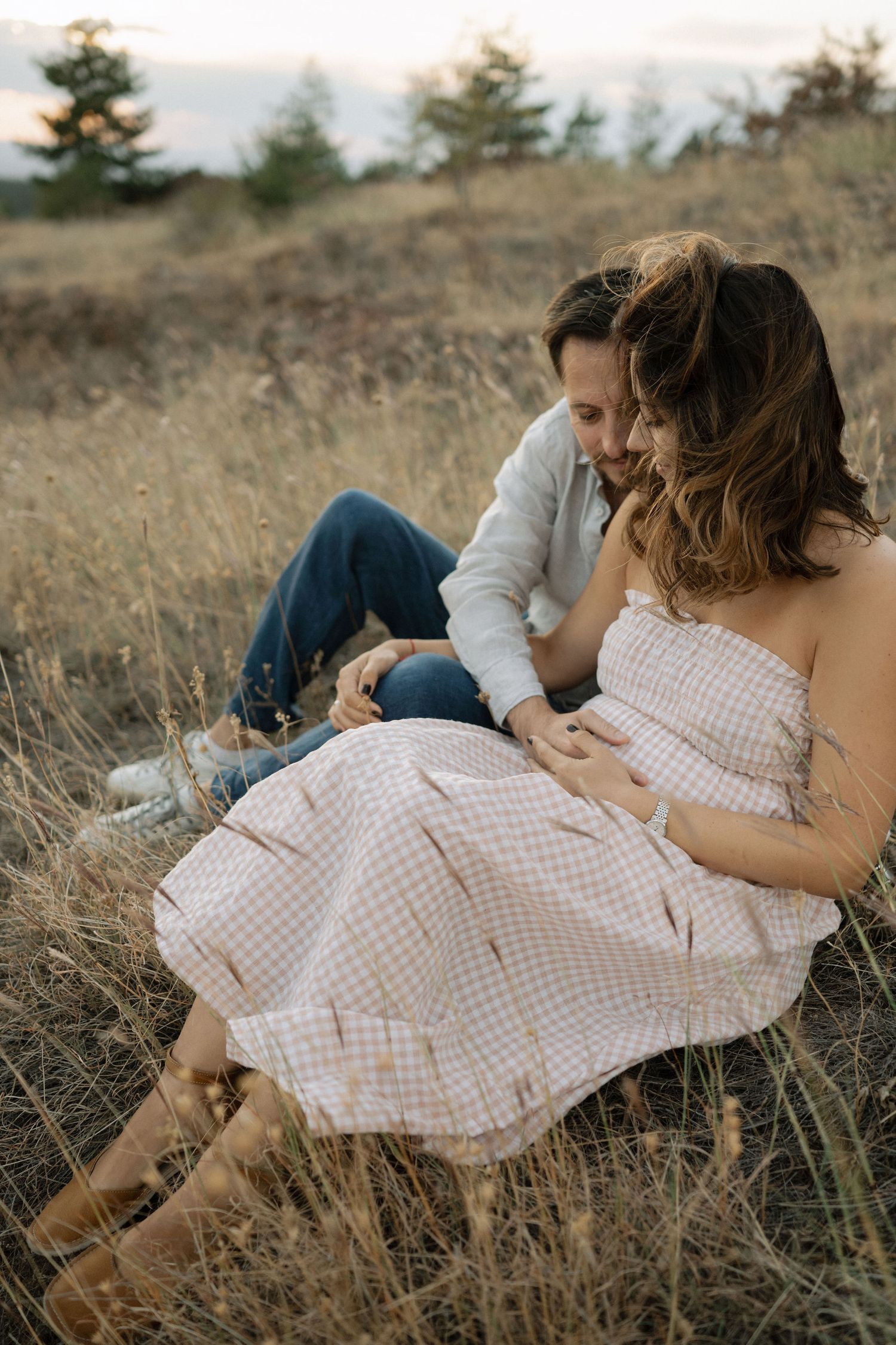 A couple in casual attire lying together on a grassy hillside during golden hour.