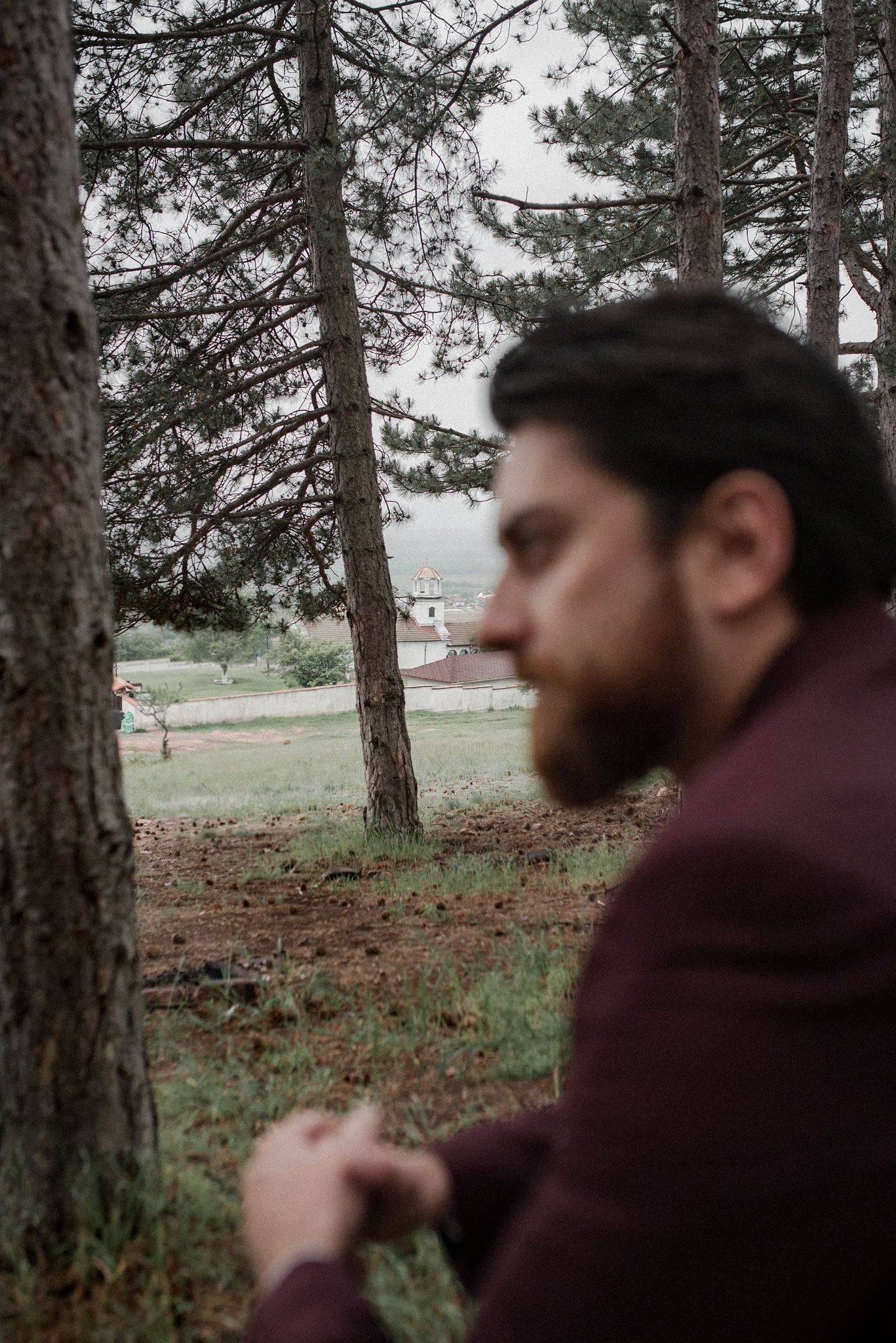 A person in a burgundy hoodie stands among tall pine trees in a misty forest setting.