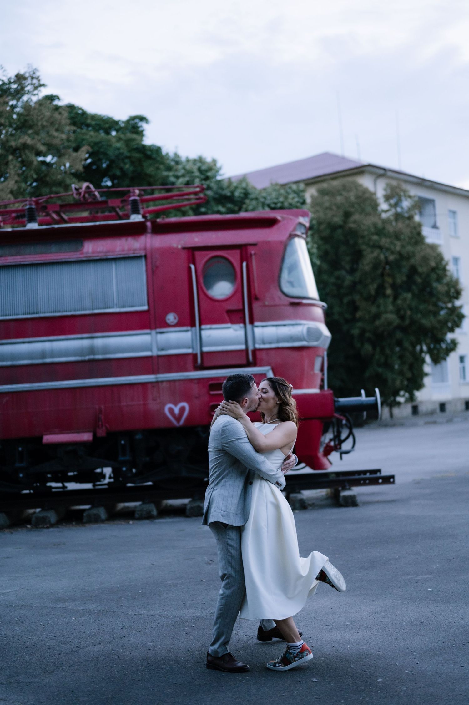 A romantic couple shares a kiss in front of a vintage red train in a dreamy outdoor setting.