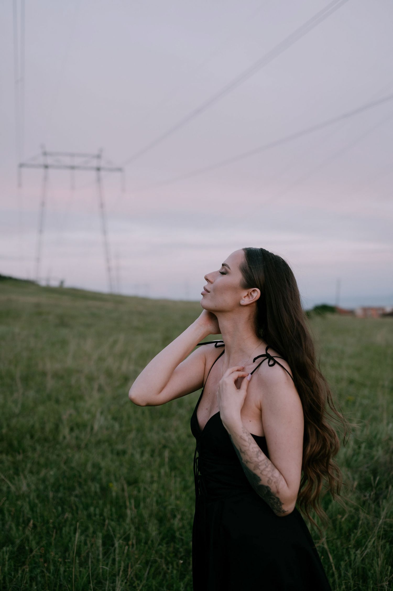 A figure in a black dress poses expressively in a grassy field at dusk with power lines visible in the background.