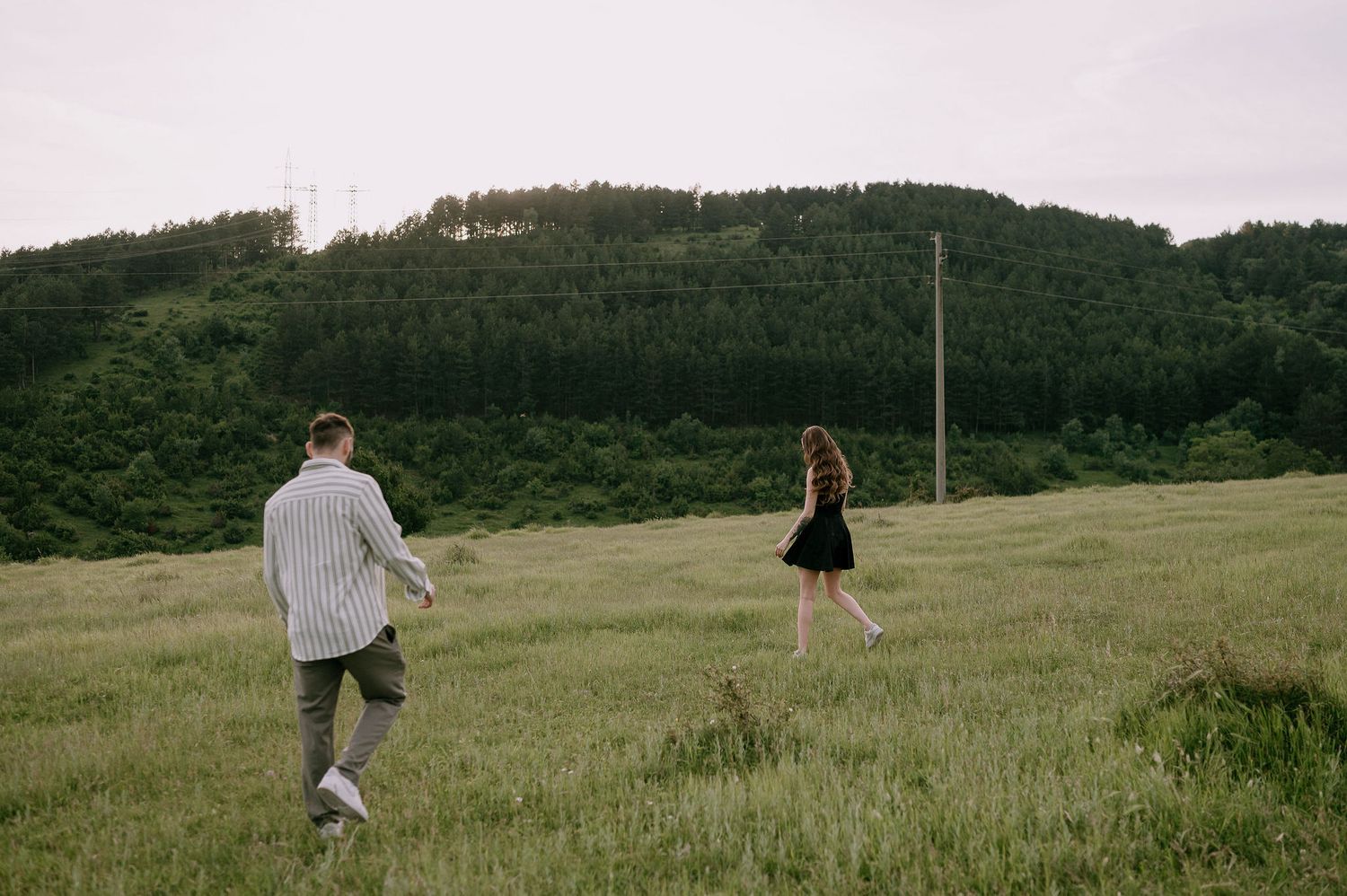 A couple walks together through a scenic green hillside meadow with forest in the background.
