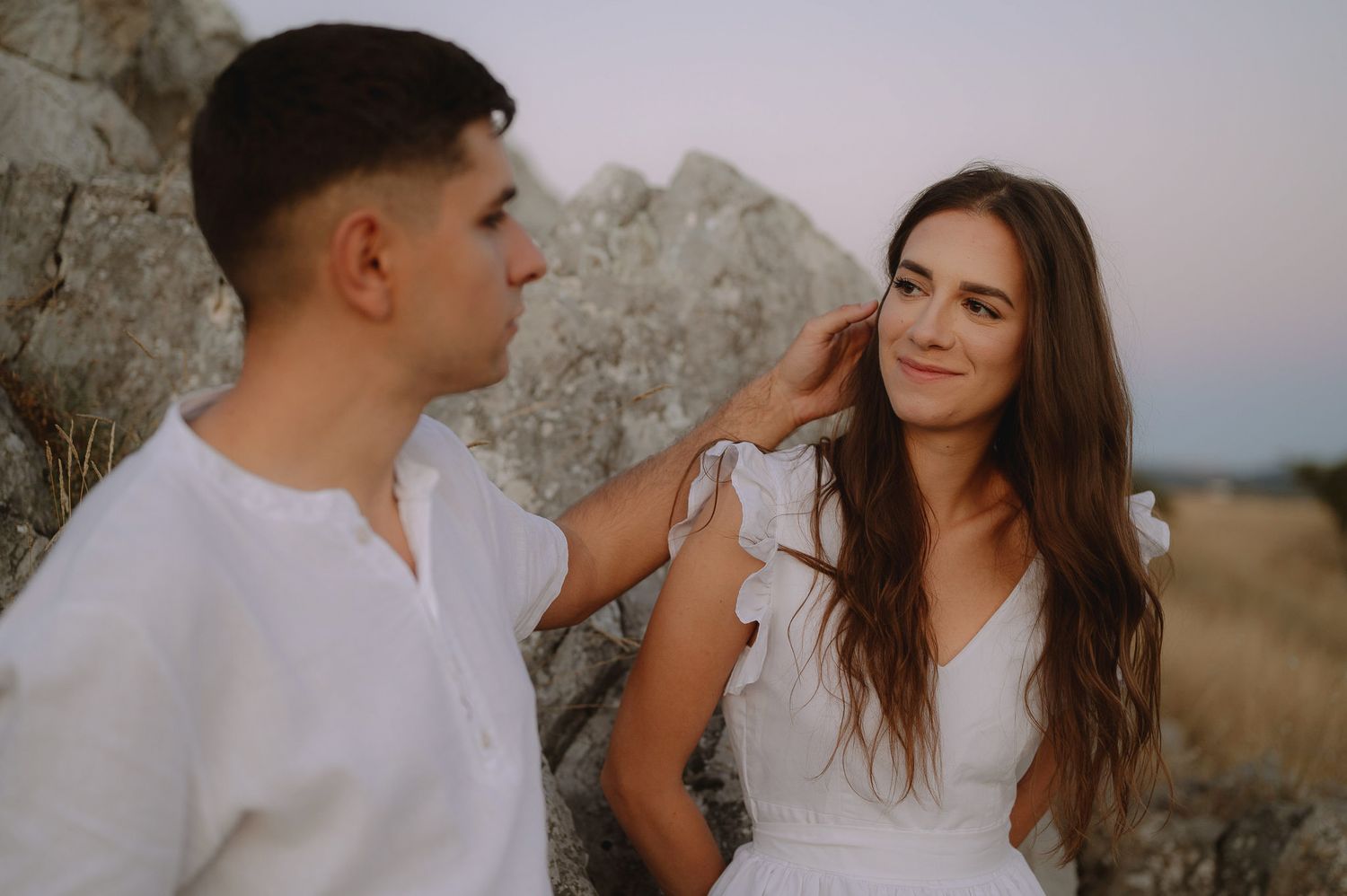 A couple in white outfits share an intimate moment against a rocky landscape at sunset.