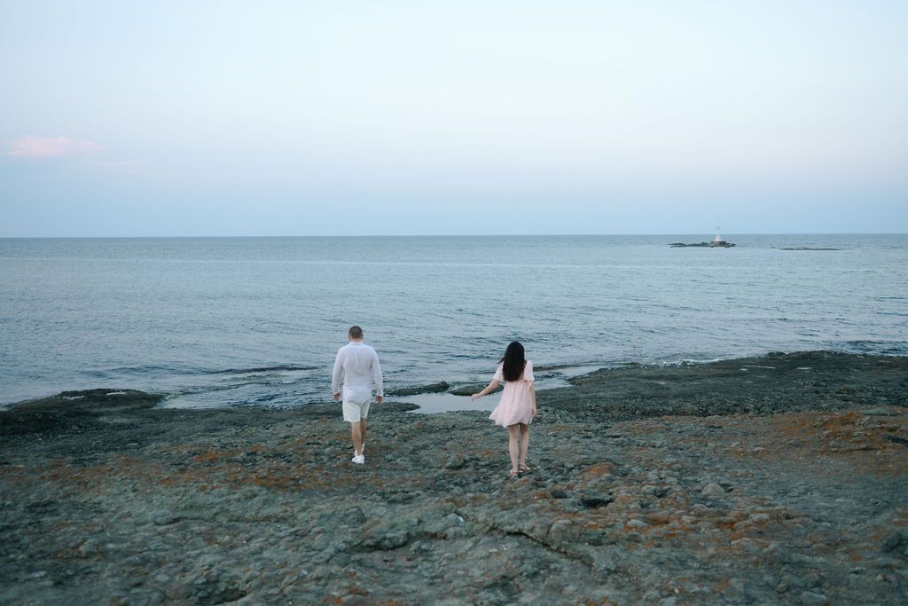 Two figures in white walk together along a rocky coastal shoreline at dusk with calm ocean waters in the background.