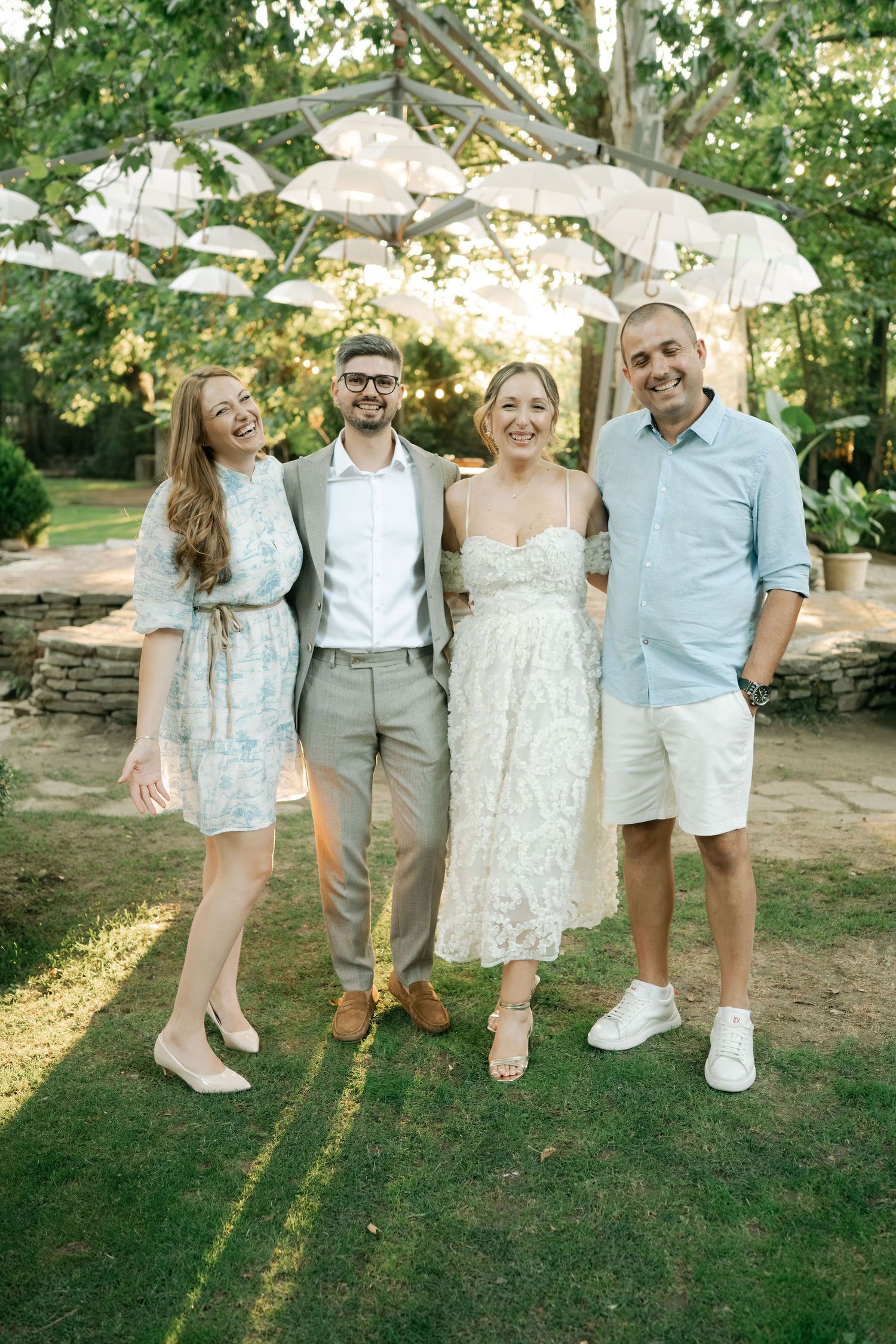 Two couples pose together outdoors in coordinated summer outfits of blue and white with parasols in the background.