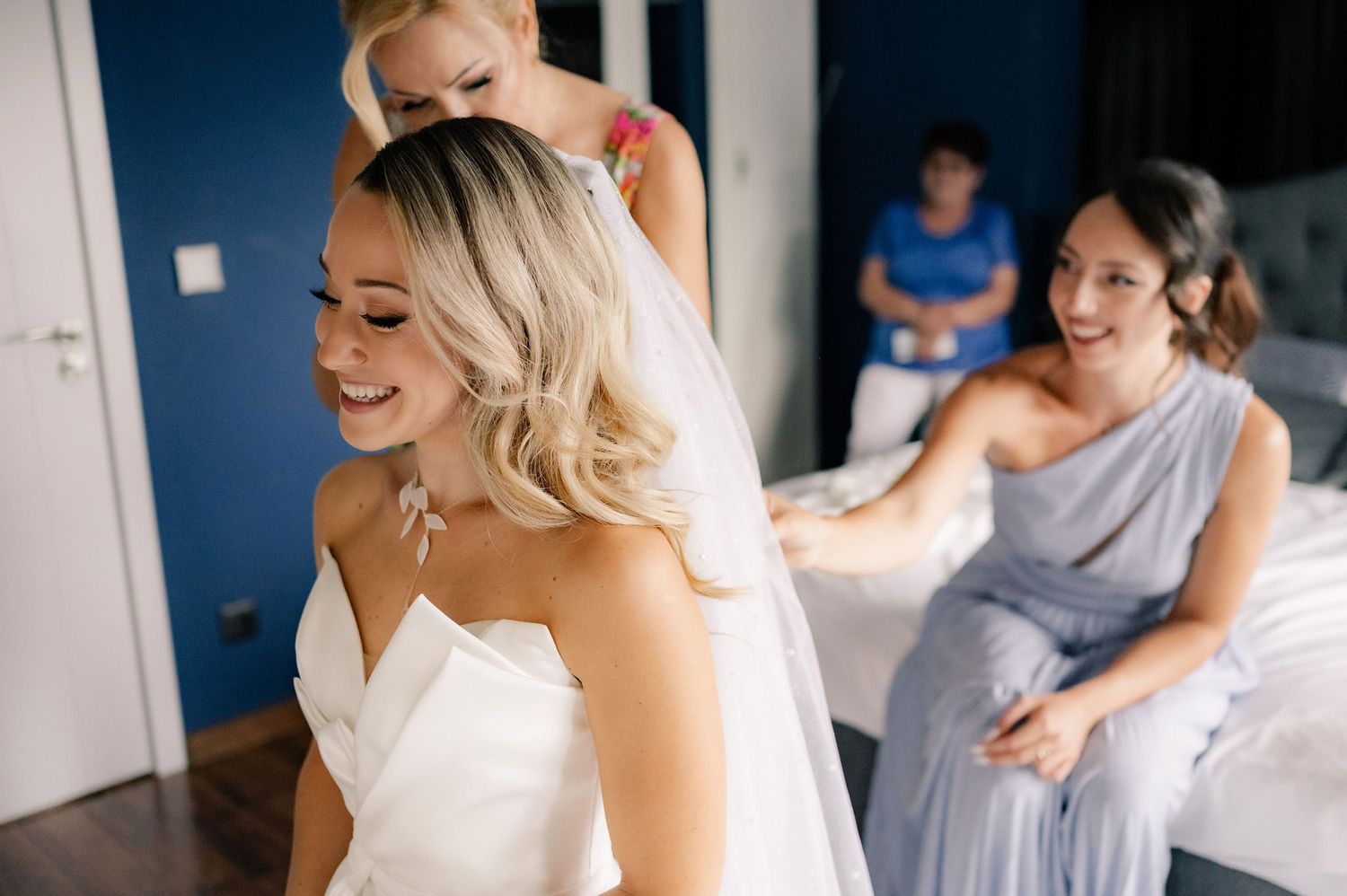A candid moment during wedding preparations shows a bride in white with bridesmaids in blue getting ready in a room.