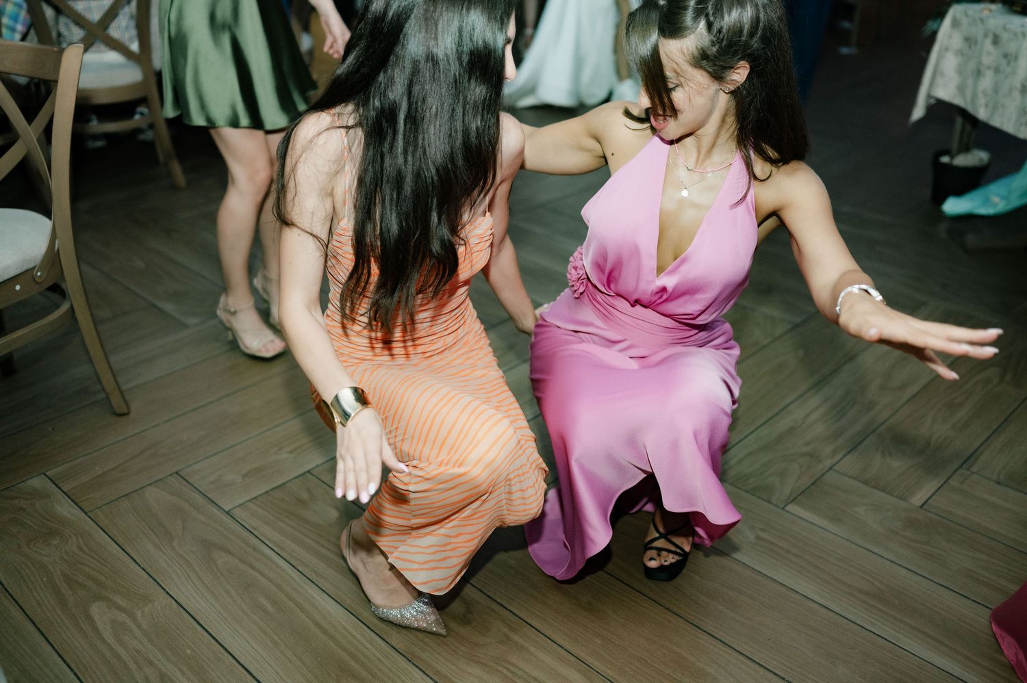 People dancing together at a party on a wooden floor in colorful dresses and casual attire.