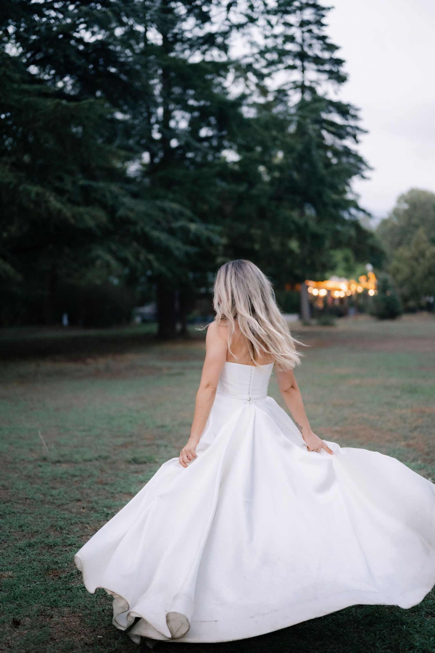 A person in a flowing white dress twirls gracefully in a grassy field at dusk with evergreen trees in the background.