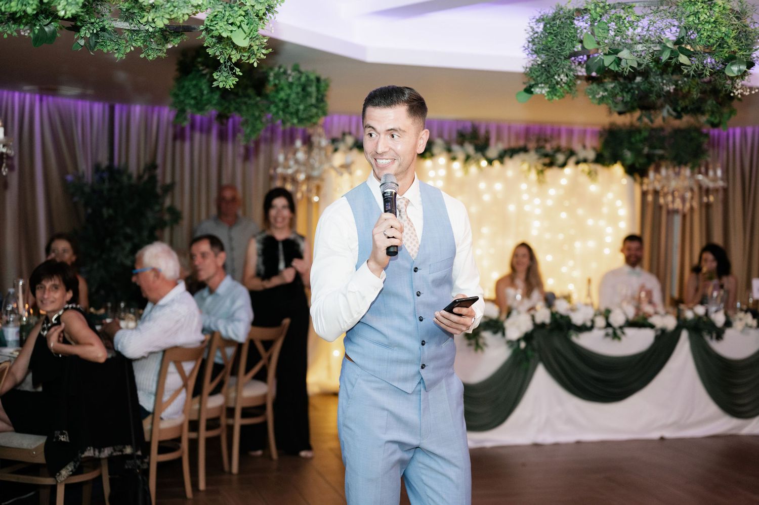 A speaker in a light gray suit addresses guests at an elegant reception venue decorated with greenery and draping.