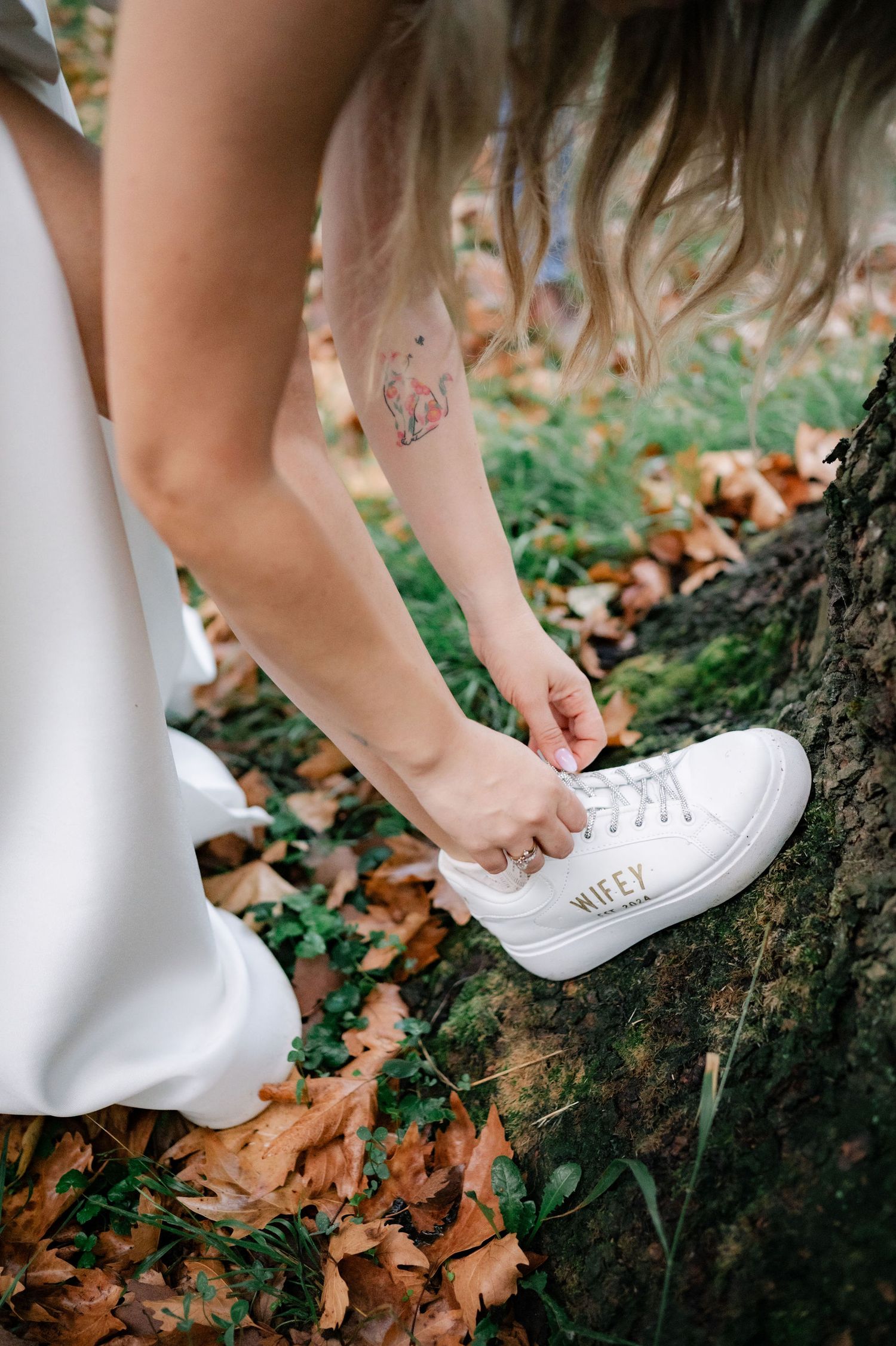 A close-up of someone adjusting white sneakers while standing on fallen autumn leaves.