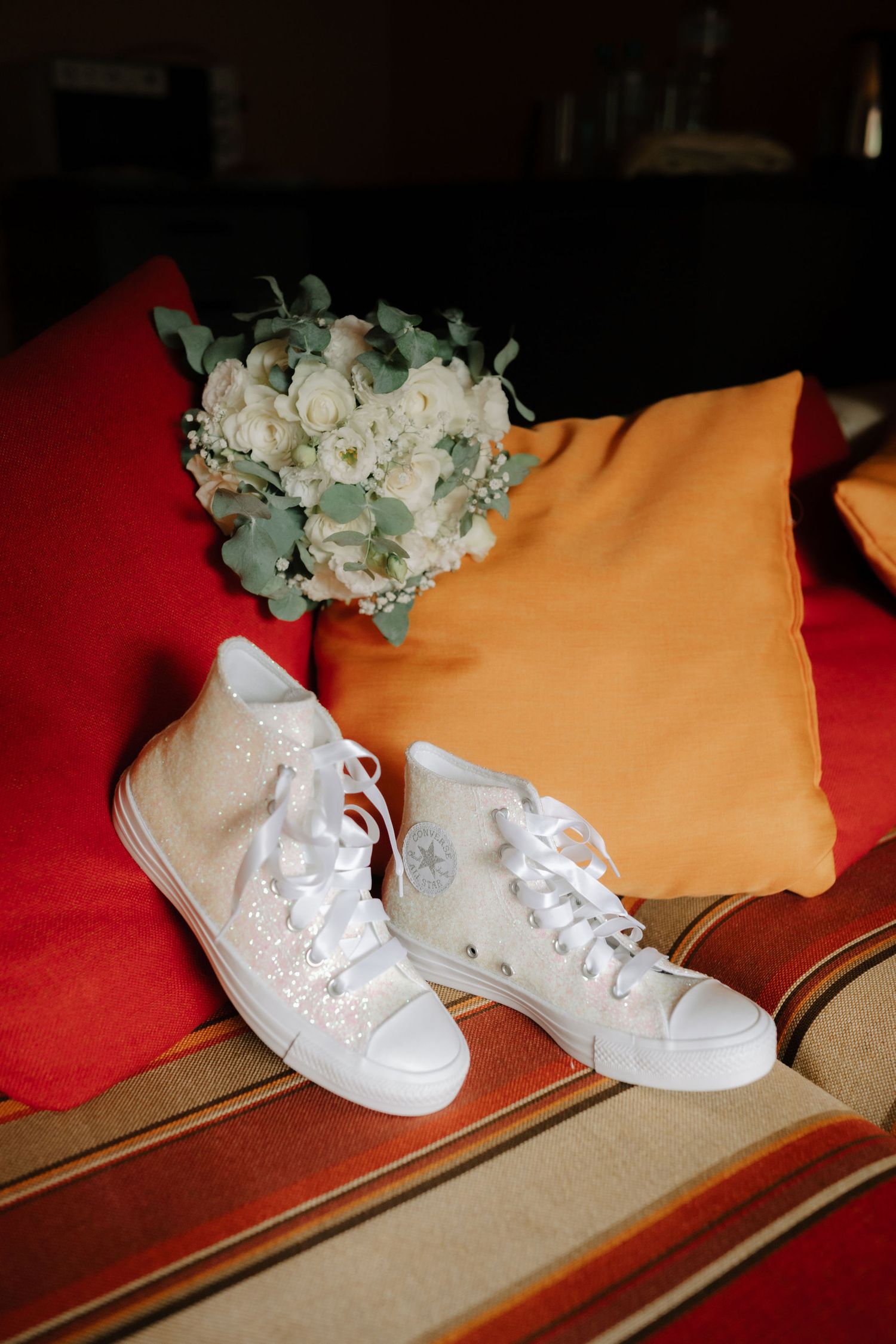 White wedding sneakers with laces sit on a red carpet next to an orange pillow and white floral bouquet.