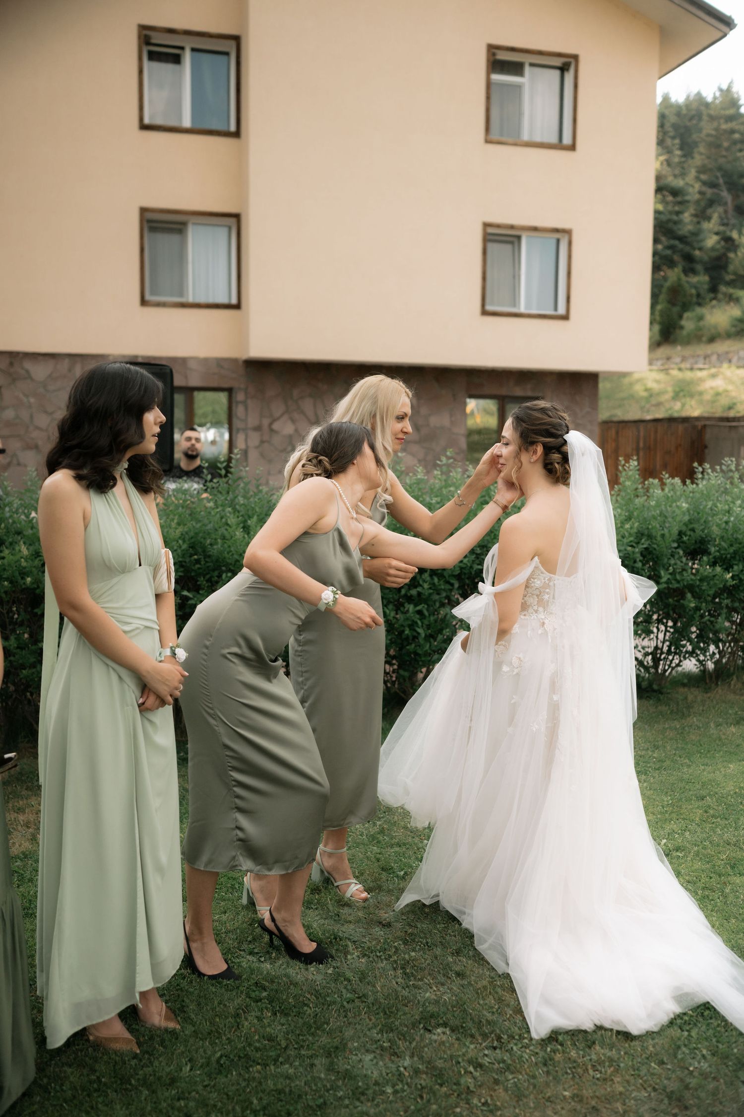 Bridesmaids in sage green dresses help adjust a bride's flowing white wedding gown outdoors.