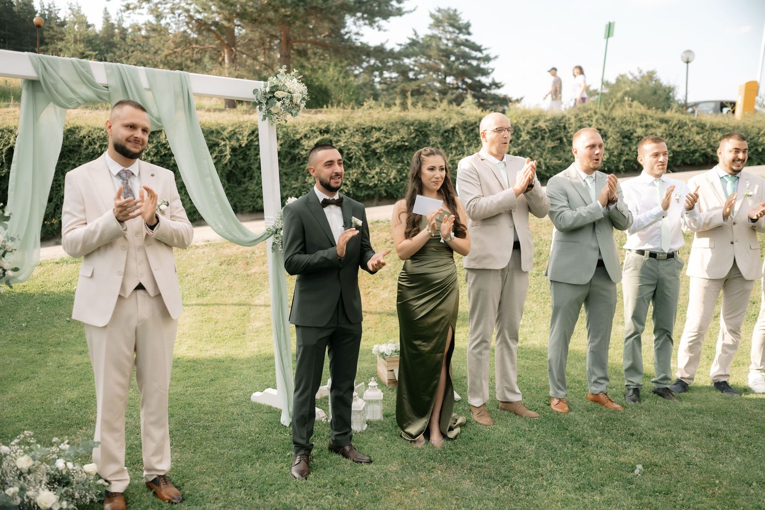 Wedding party members stand in a row on green grass wearing coordinated beige and gray formal attire.