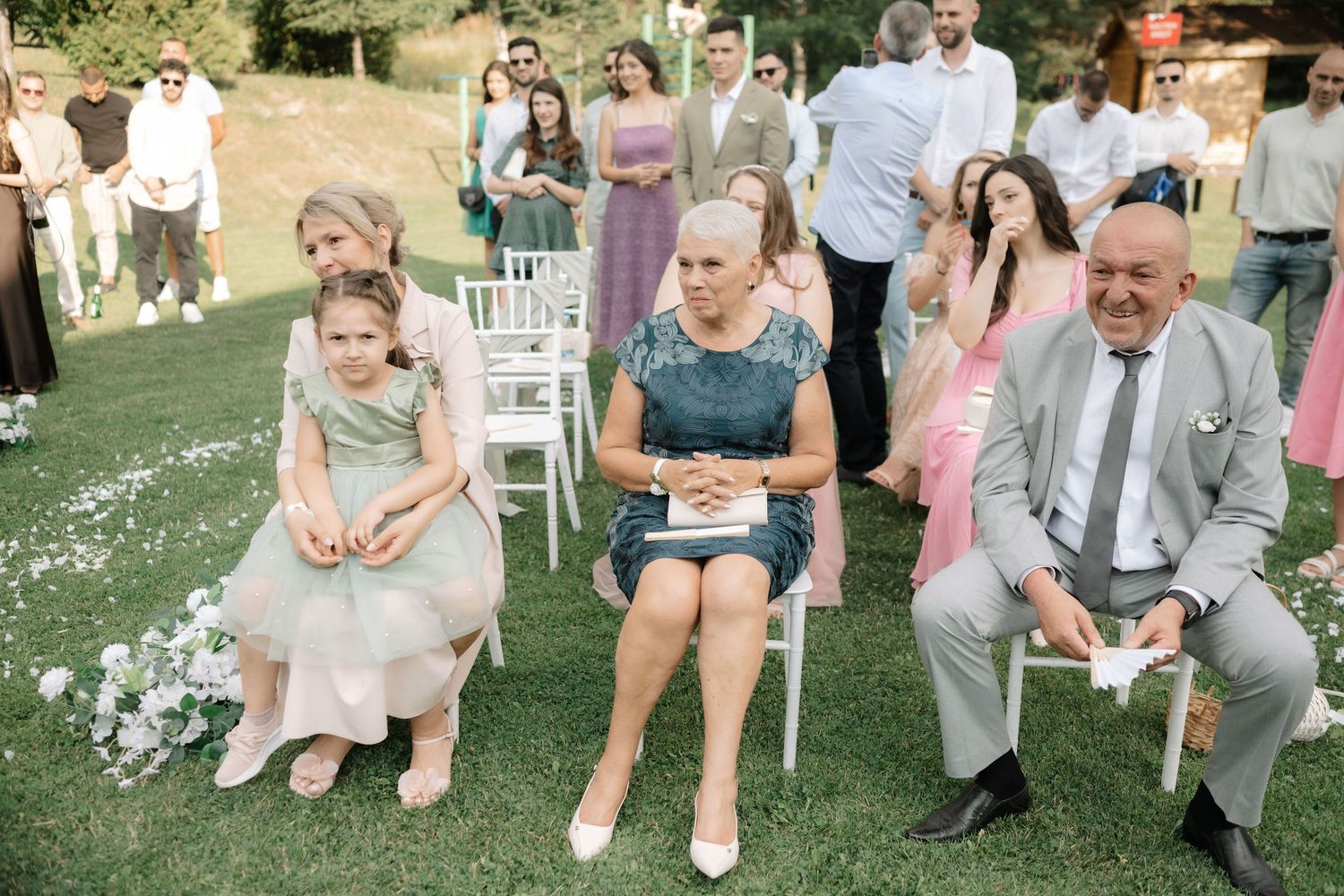 Guests sitting in white chairs on a lawn during an outdoor wedding ceremony with scattered flower petals on the grass.
