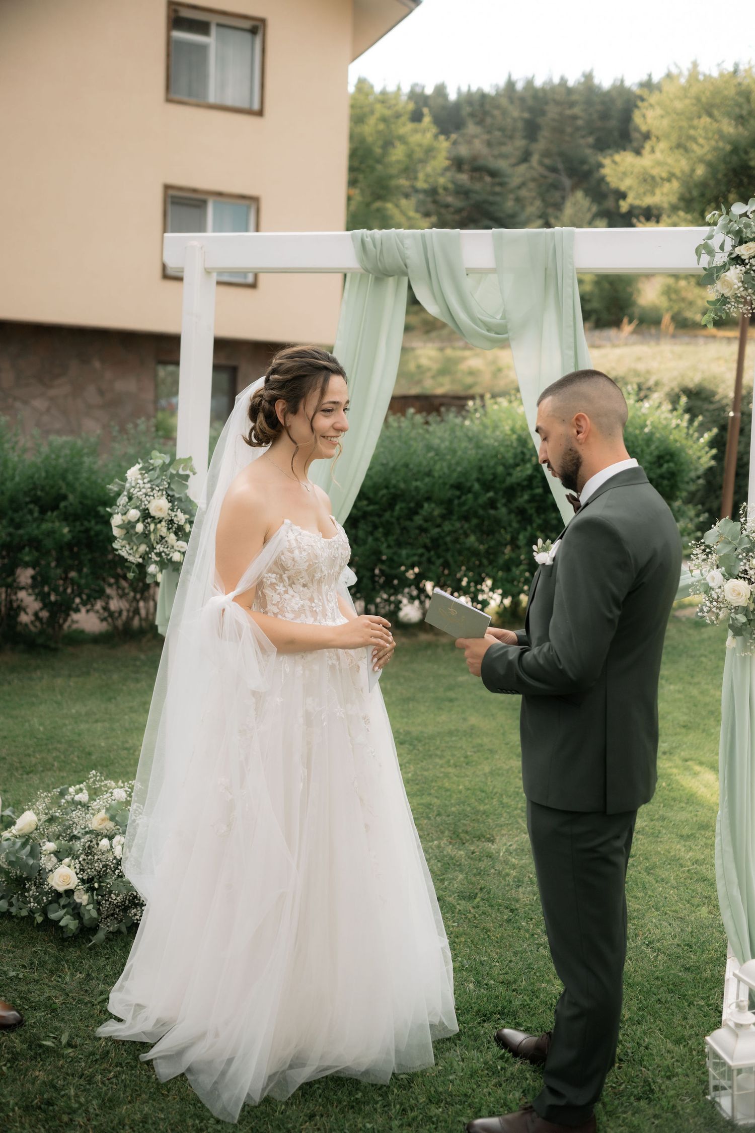 A romantic first look moment at an outdoor wedding ceremony under a white draped arch with lush greenery.