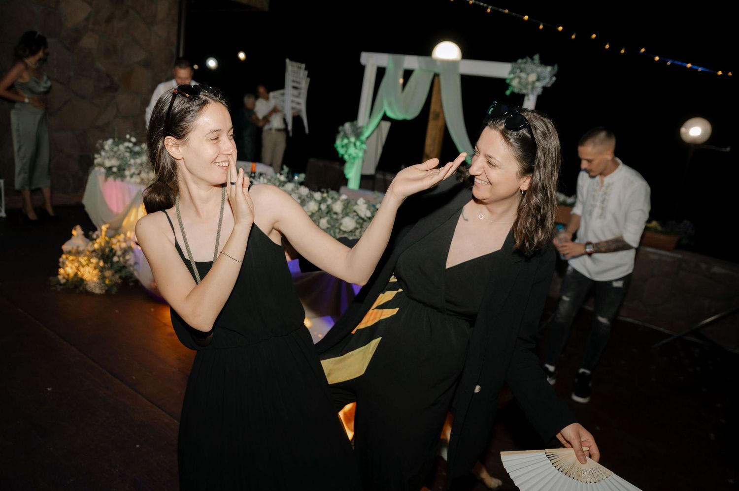 Two people in black dresses dancing together at a nighttime party with string lights overhead.