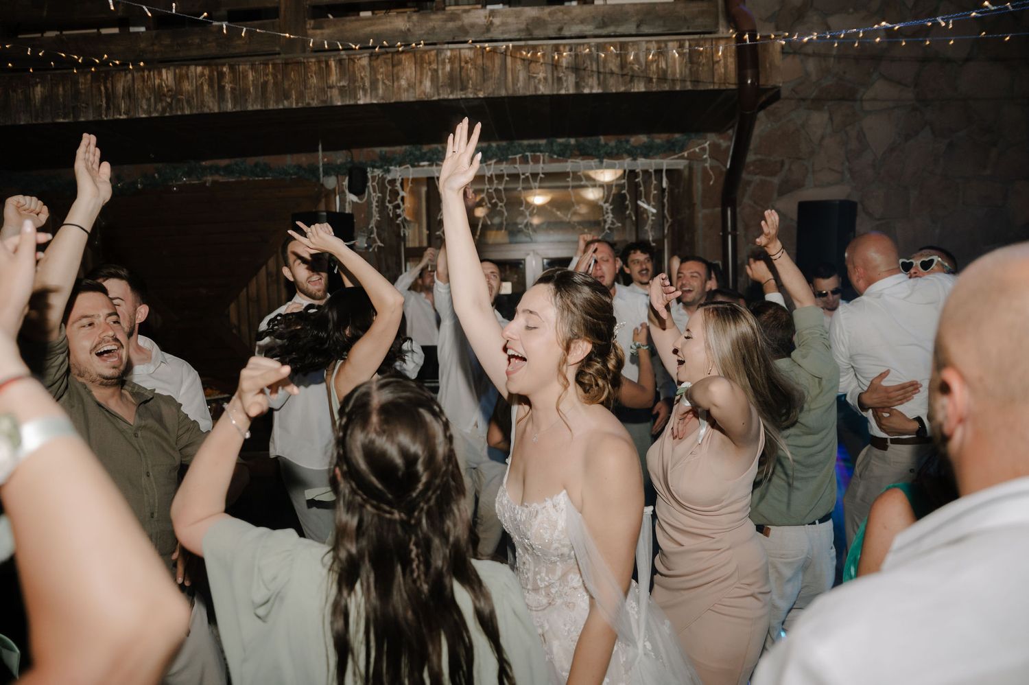 Wedding guests dance and celebrate with raised arms on a dimly lit dance floor during a reception party.