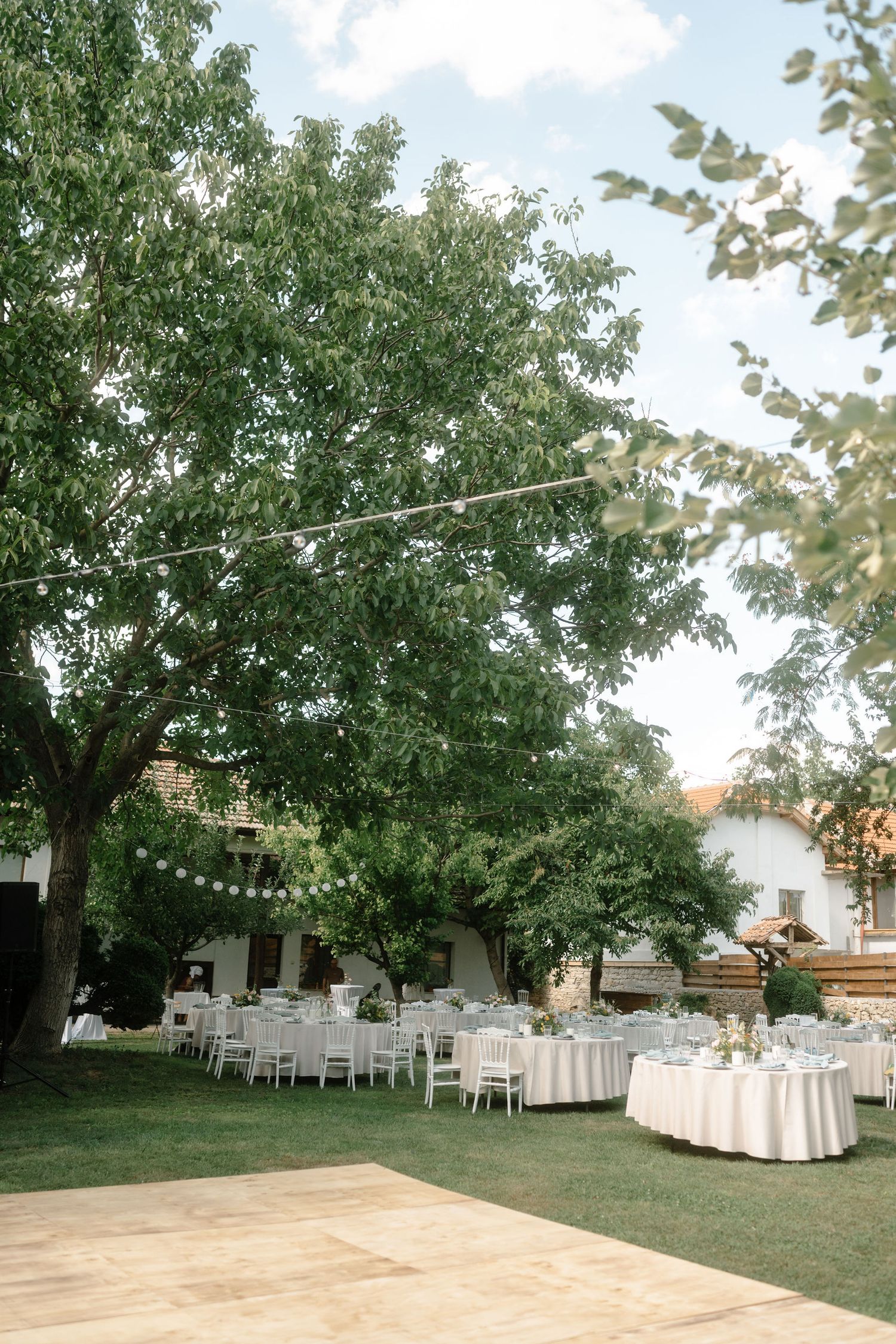 String lights hang between trees over an outdoor wedding reception area with white tables and a wooden dance floor.