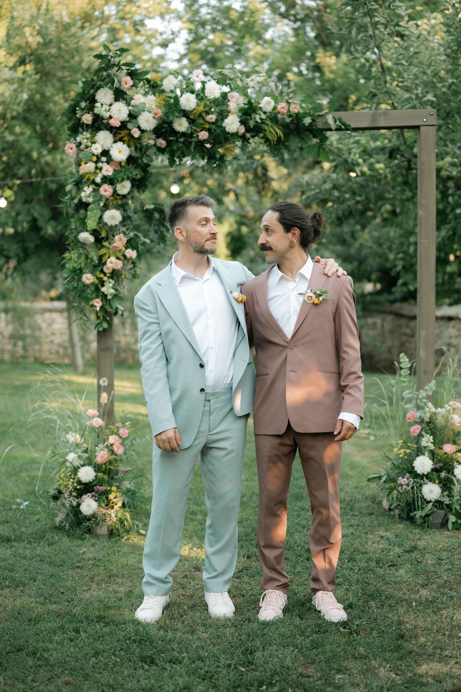 Two people in coordinating suits stand together on grass with blooming rose bushes in the background.