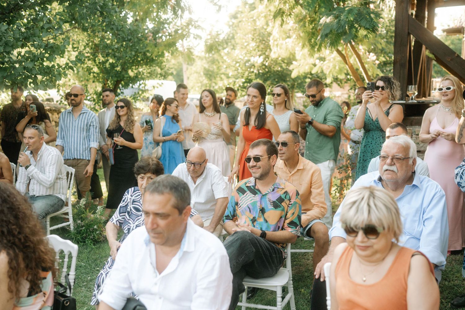 Wedding guests gather outdoors in a garden setting during a sunny afternoon celebration.