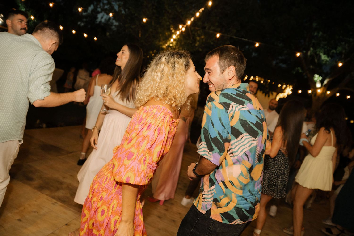 People dancing and celebrating at a lively outdoor nighttime party with string lights overhead.