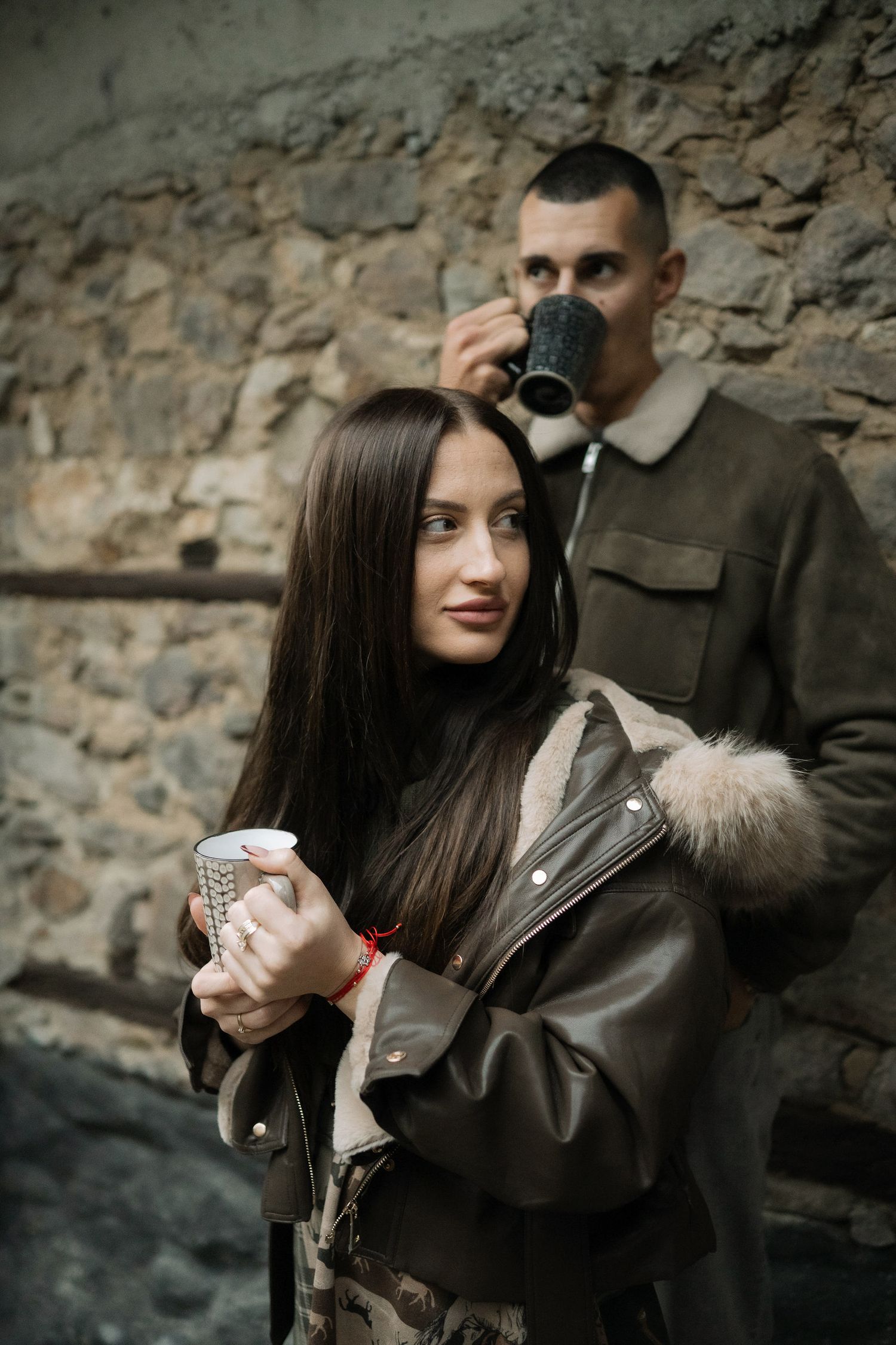 Person in leather jacket holding a beverage while standing against stone wall in moody lighting.
