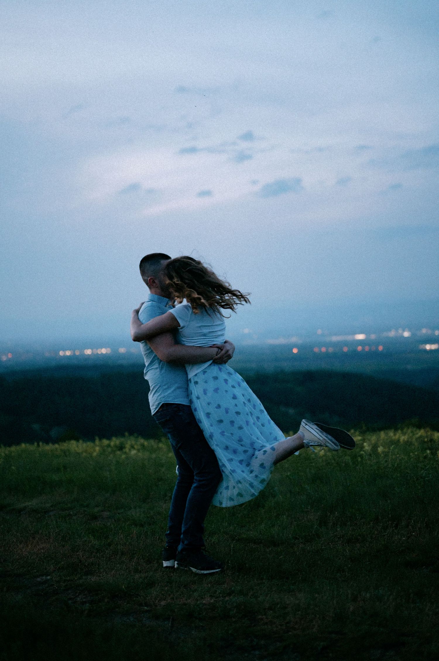 Two people share a romantic dance on a grassy hilltop overlooking city lights at twilight.