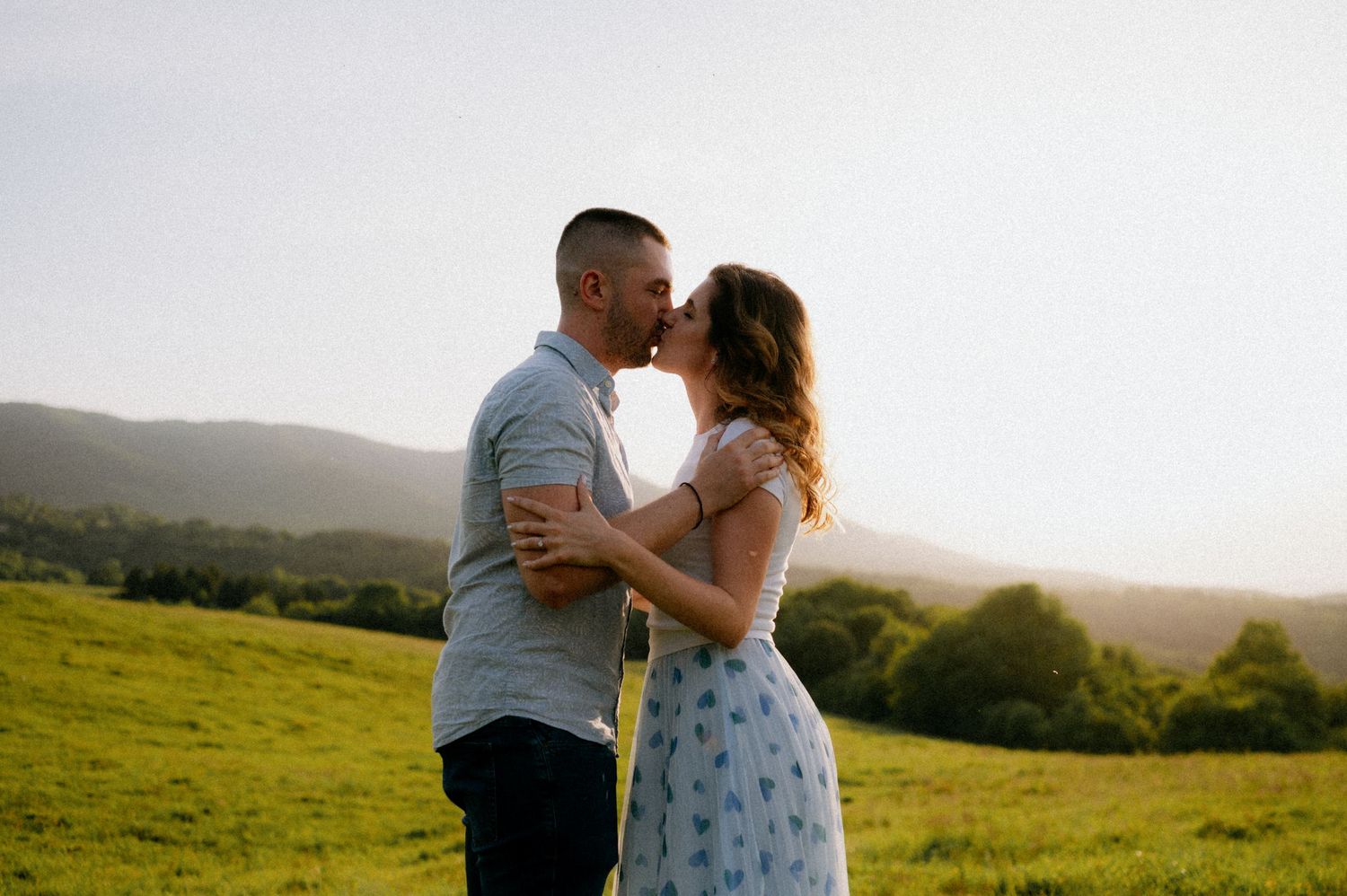 Couple embracing during sunset photoshoot in a scenic hillside meadow.