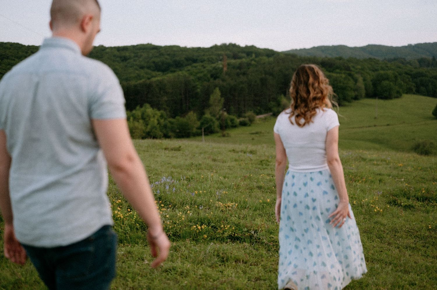 Two people walk hand in hand through a lush green hillside with mountains in the distance.