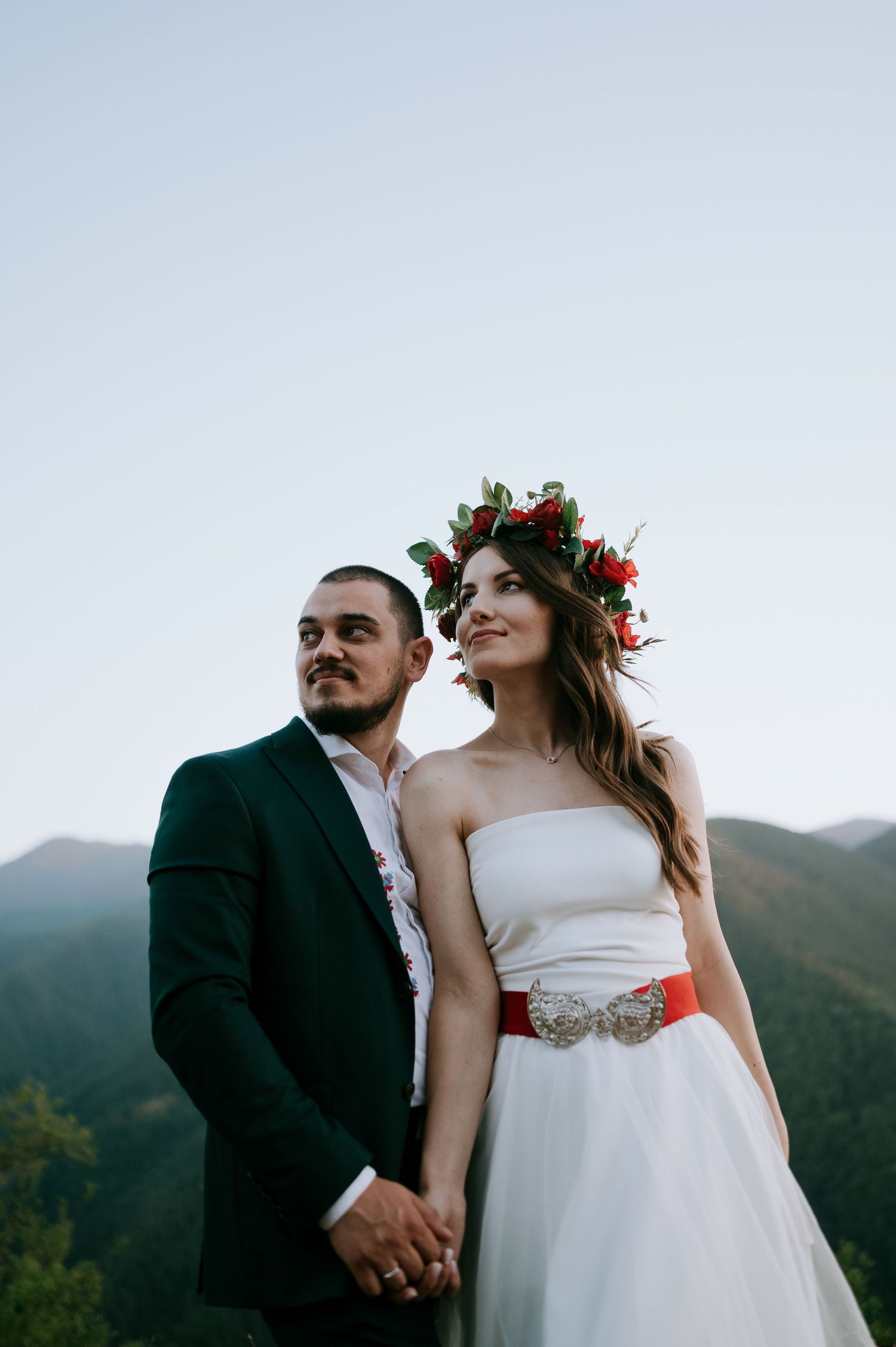 A couple in wedding attire stands together on a mountain overlook at dusk.