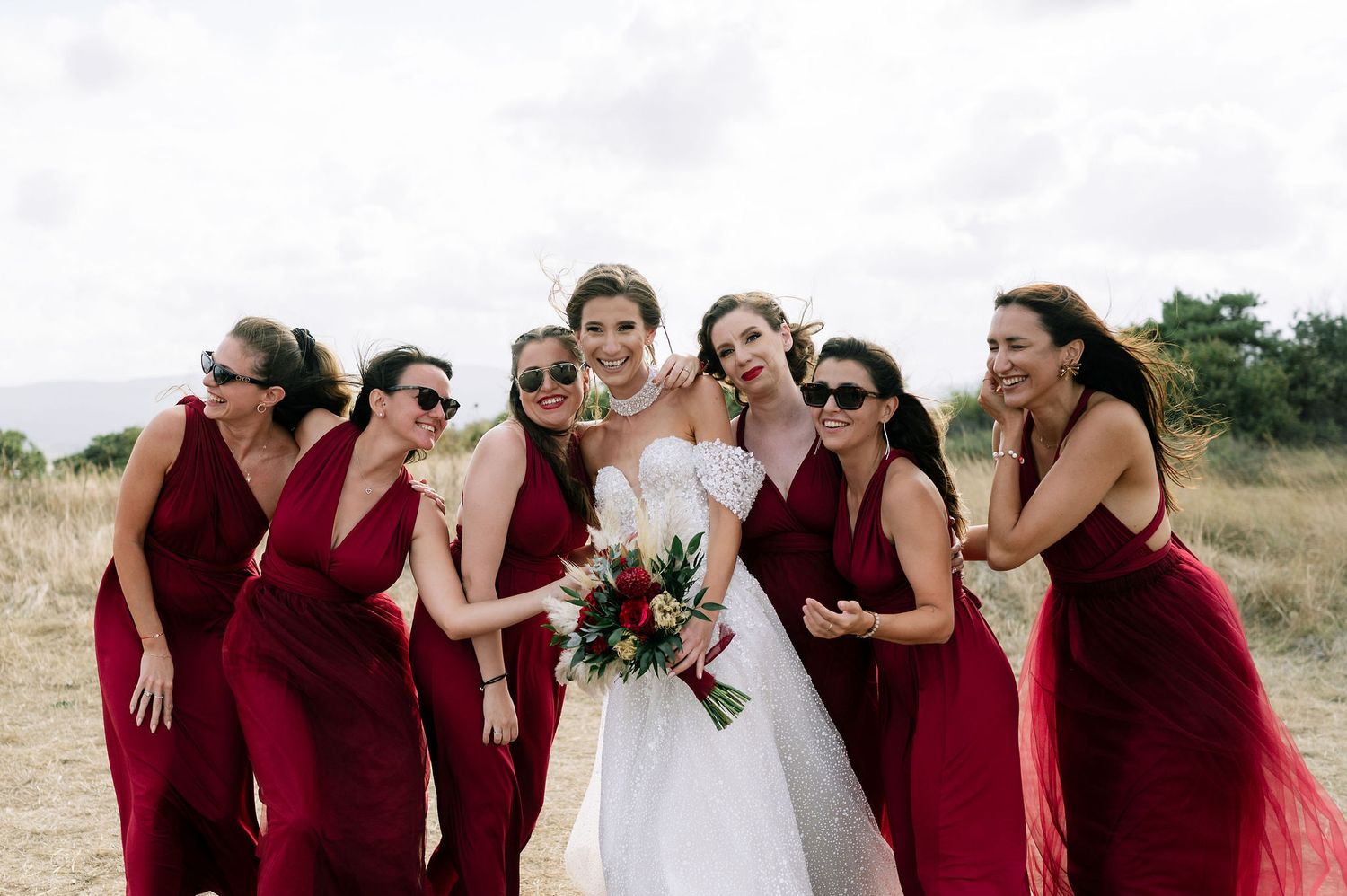 Wedding group in burgundy dresses and white gown share a joyful moment outdoors.