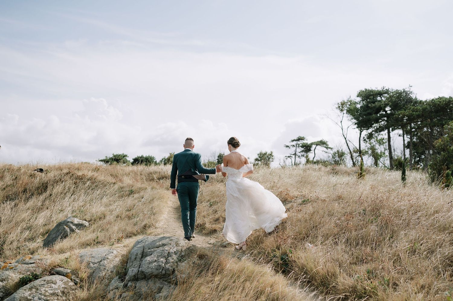 Couple in wedding attire strolls along scenic hilltop path with ocean views.