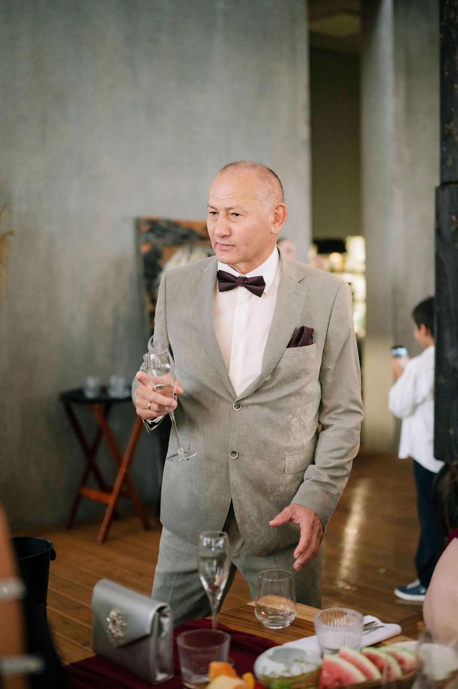 Guest in gray suit and bowtie holds champagne glass at formal indoor event.