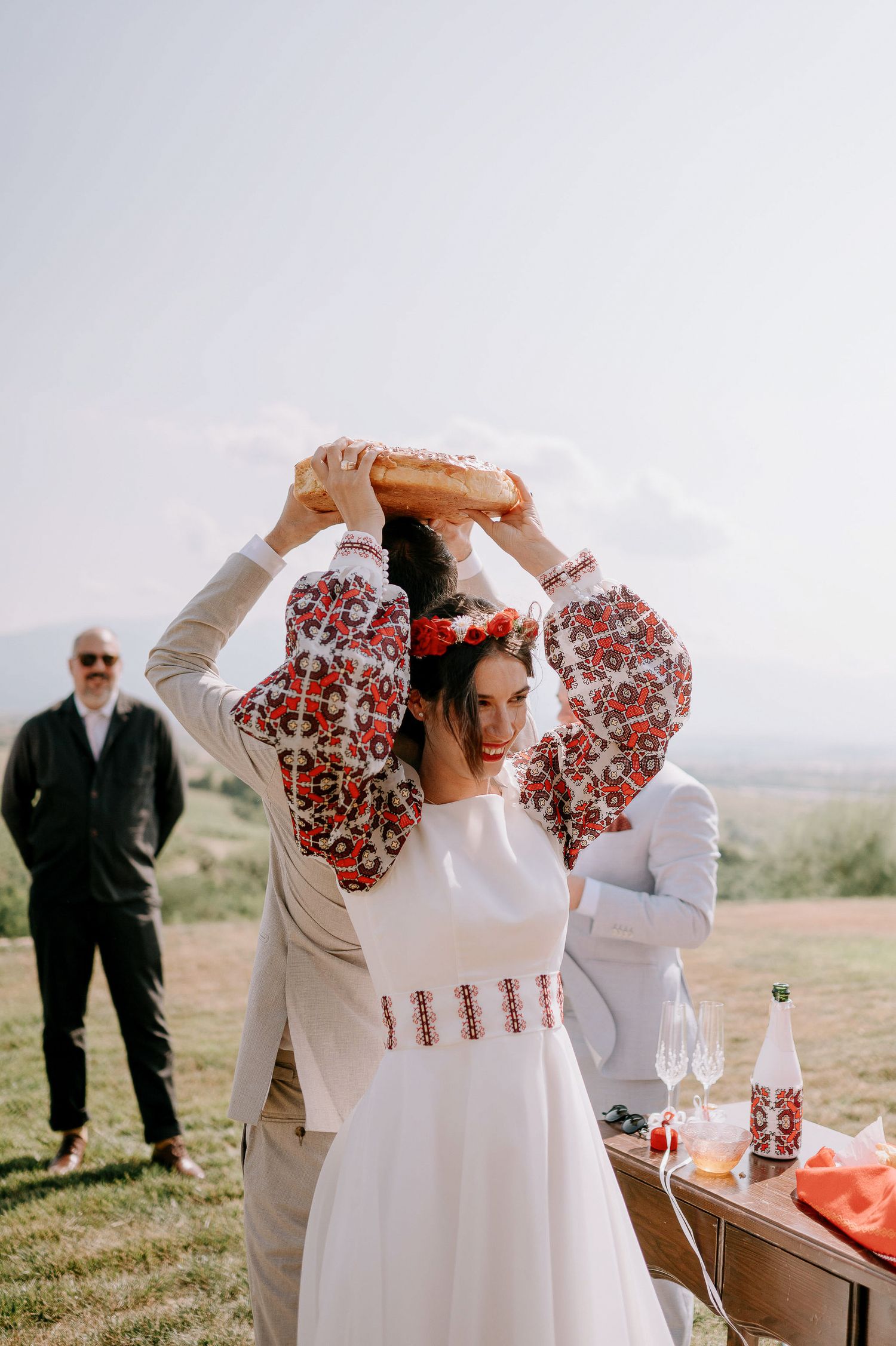 Wedding ceremony participants in traditional white dresses and light suits gather on a hilltop.