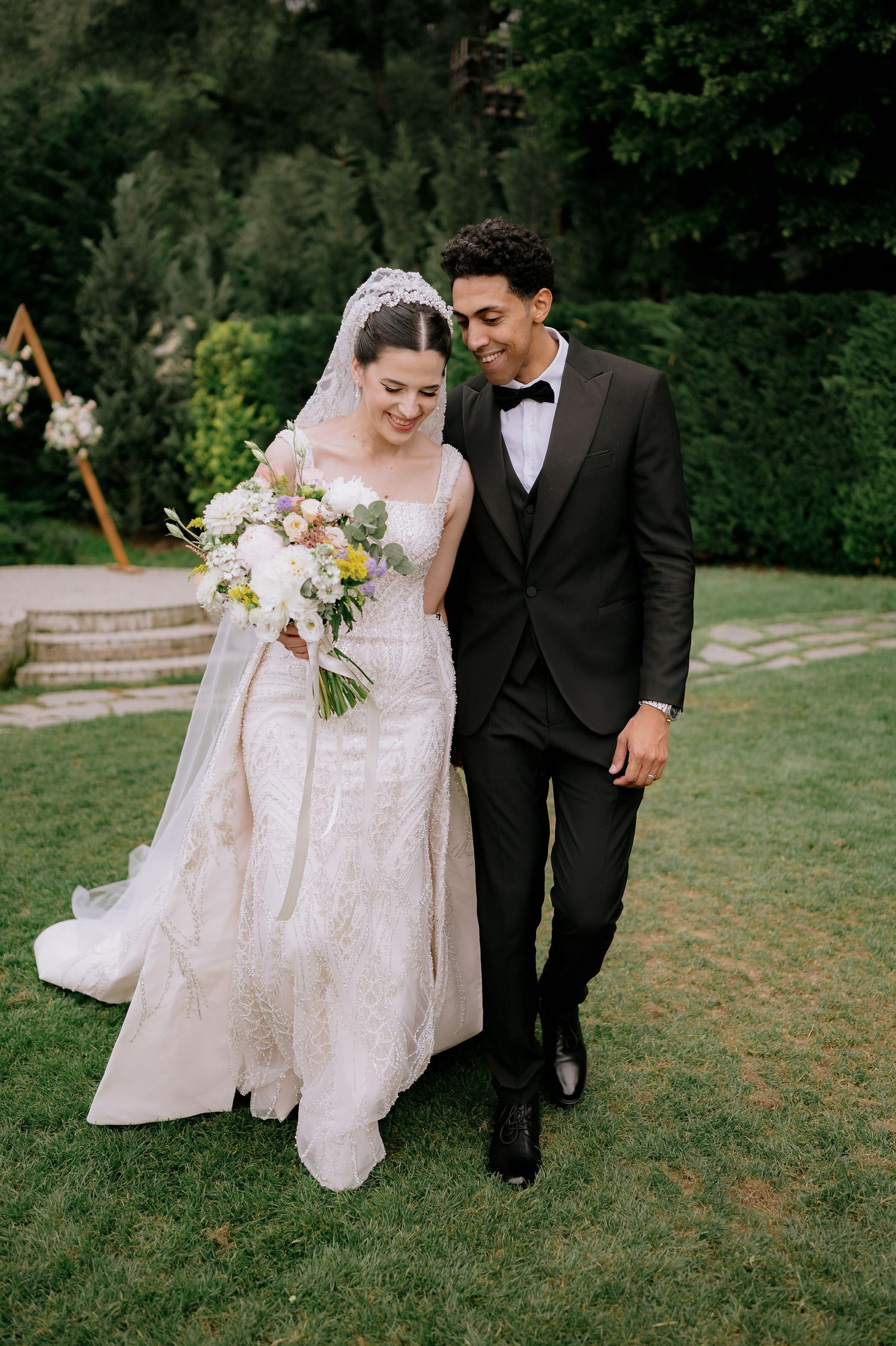 A couple walking together on a grassy lawn, with the bride wearing a flowing white dress and groom in black tuxedo.