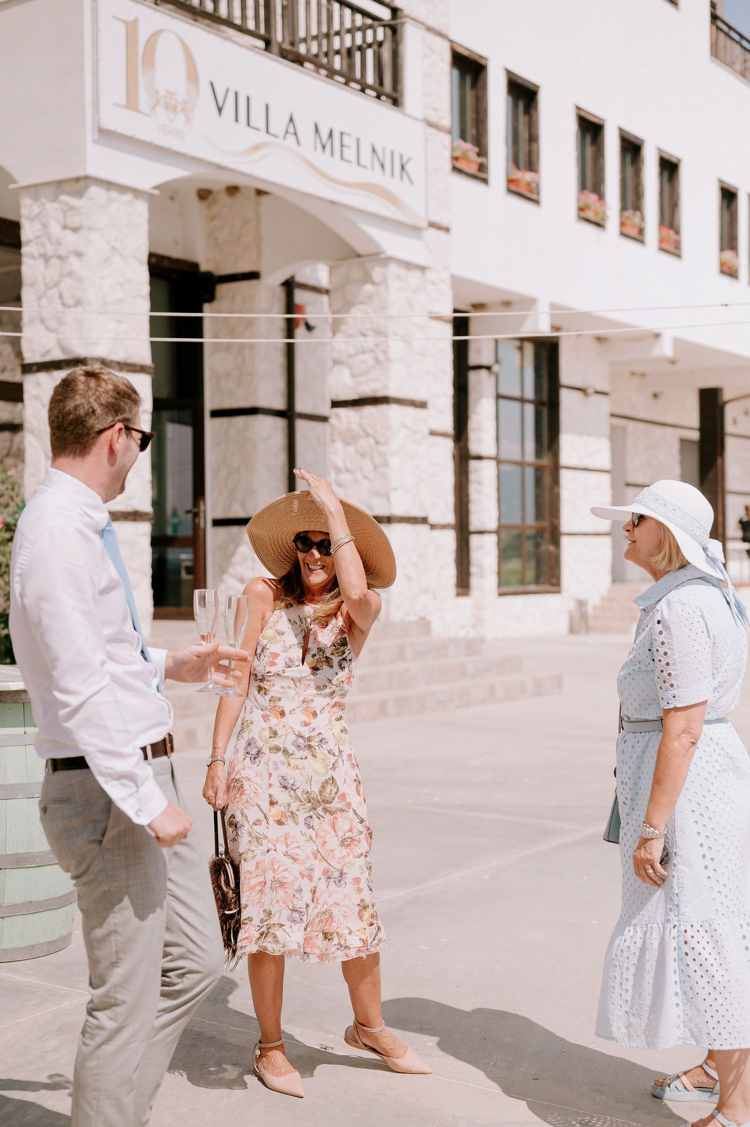 People in summer attire walking and chatting outside Villa Melnik's white stone building.